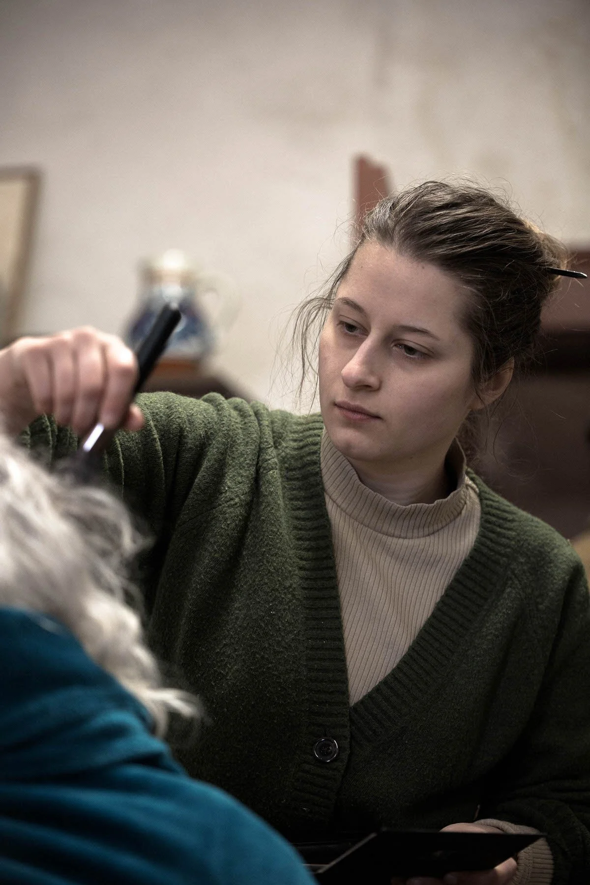 Une jeune femme avec des cheveux bruns attachés, portant un pull vert et un t-shirt beige, maquillant un acteur pour une scène