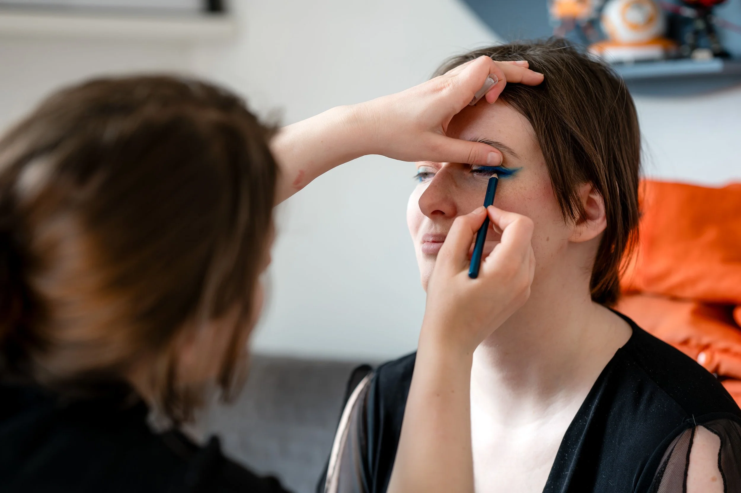 Une femme applique du maquillage bleu autour de l'œil d'une autre femme, qui se tient debout, dans un intérieur domestique.