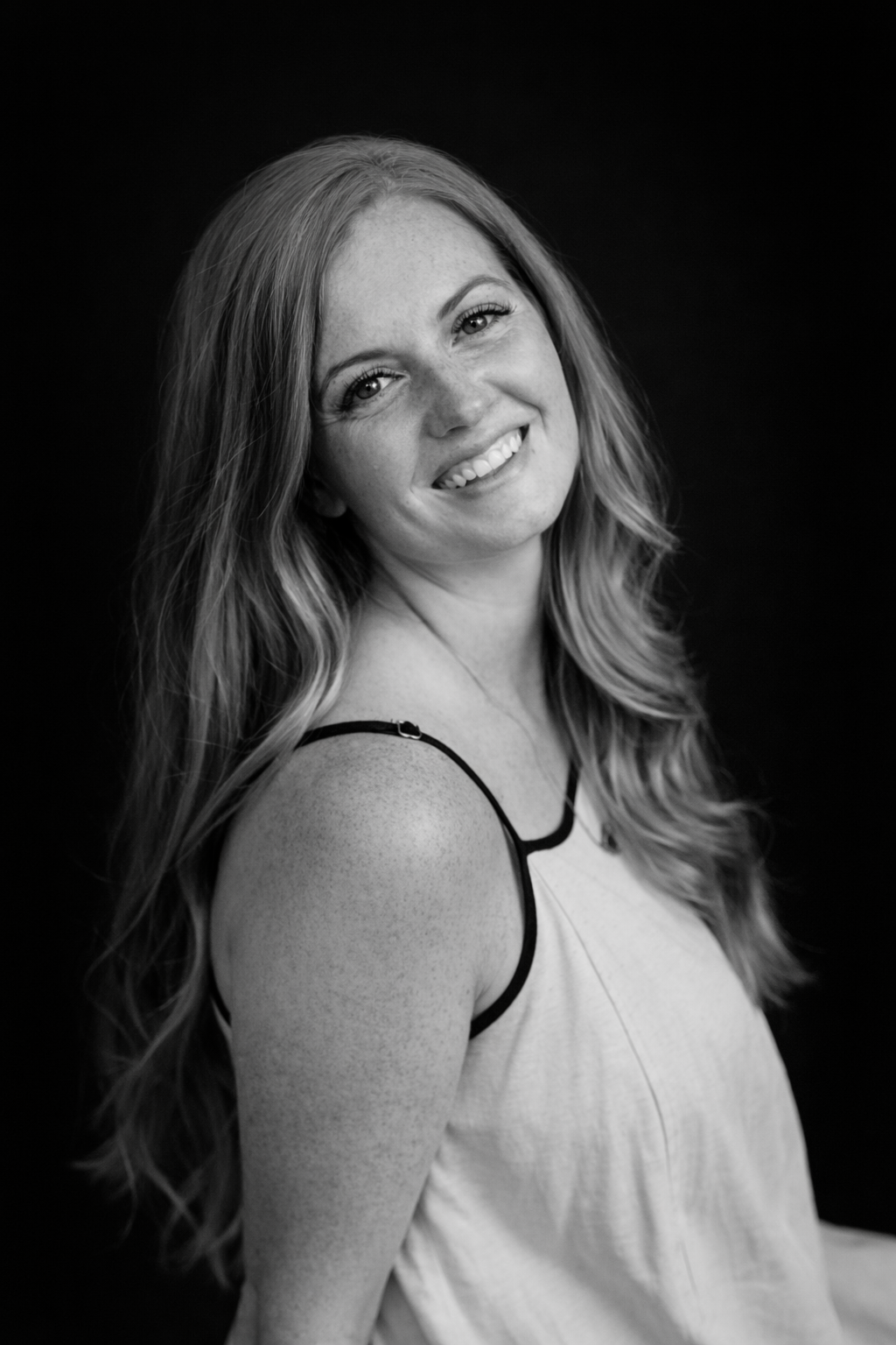 Black and white portrait of a smiling young woman with long wavy hair, wearing a sleeveless top with thin straps, against a dark background.