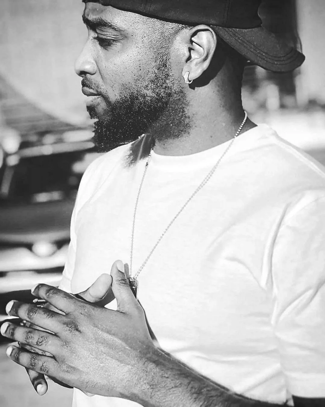 Black and white photo of a man wearing a backward cap, a white t-shirt, and a necklace. He has a beard and is holding his hands together. The background is blurred.