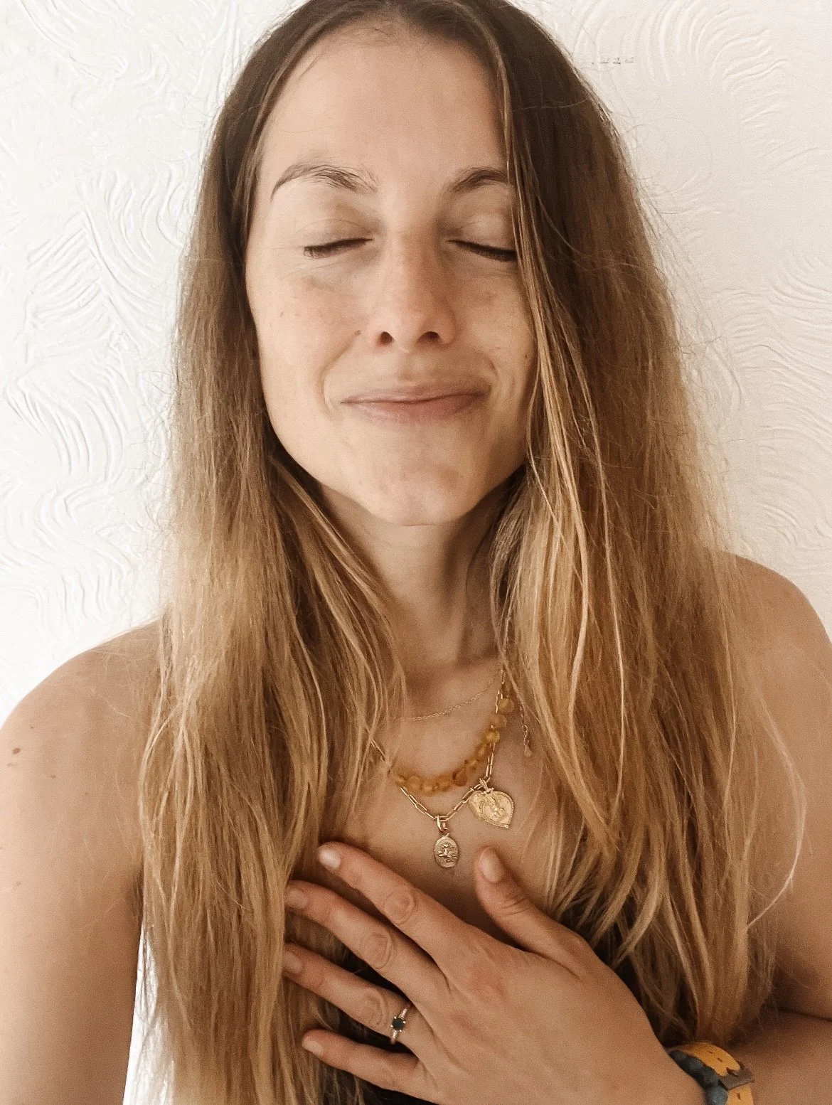 Woman with long wavy hair and gold jewelry, eyes closed, hand on chest, standing against white textured wall.