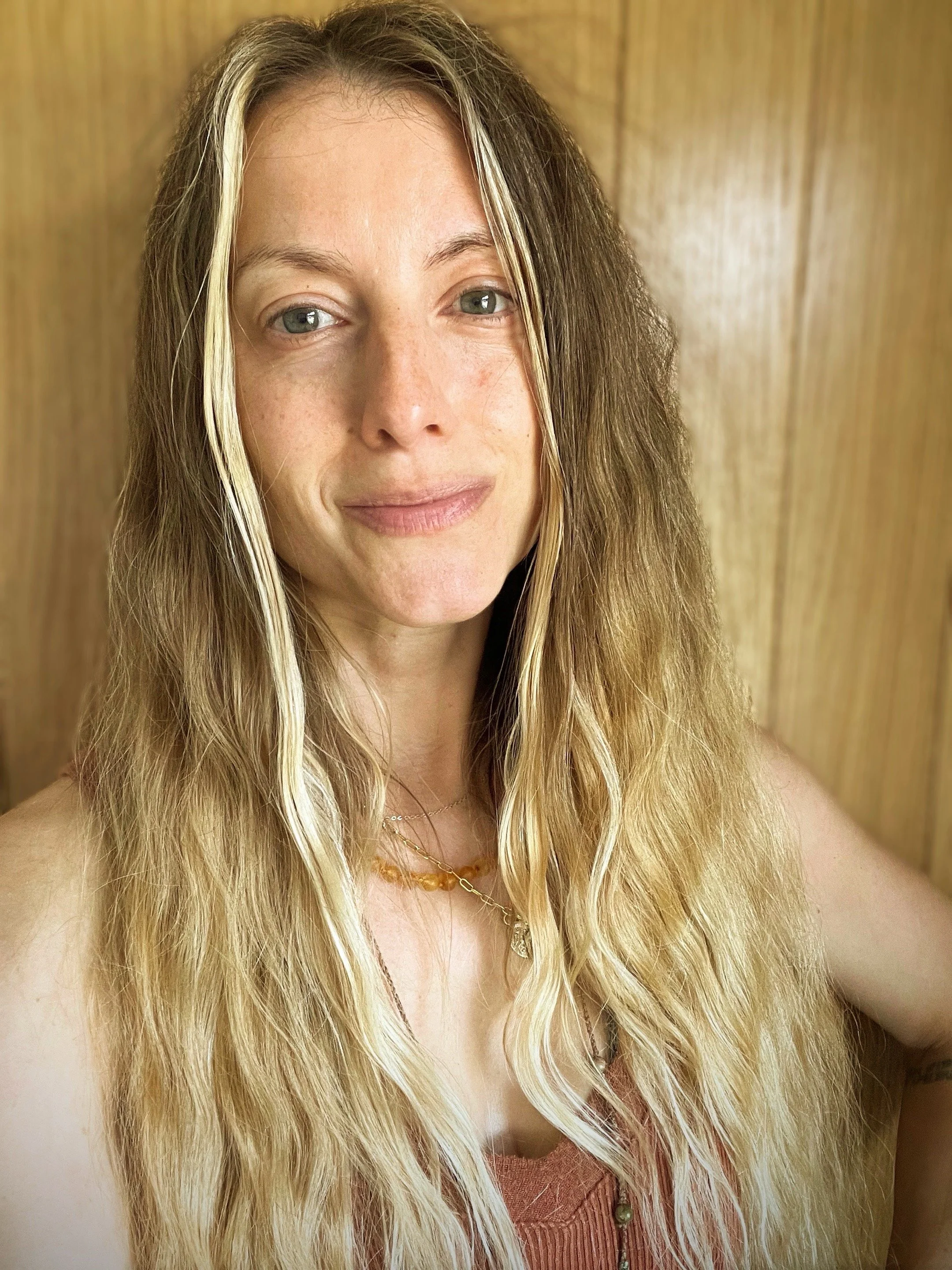 A woman with long, wavy blonde hair and light skin, smiling softly, standing in front of a wooden wall, wearing a tan top and necklaces.
