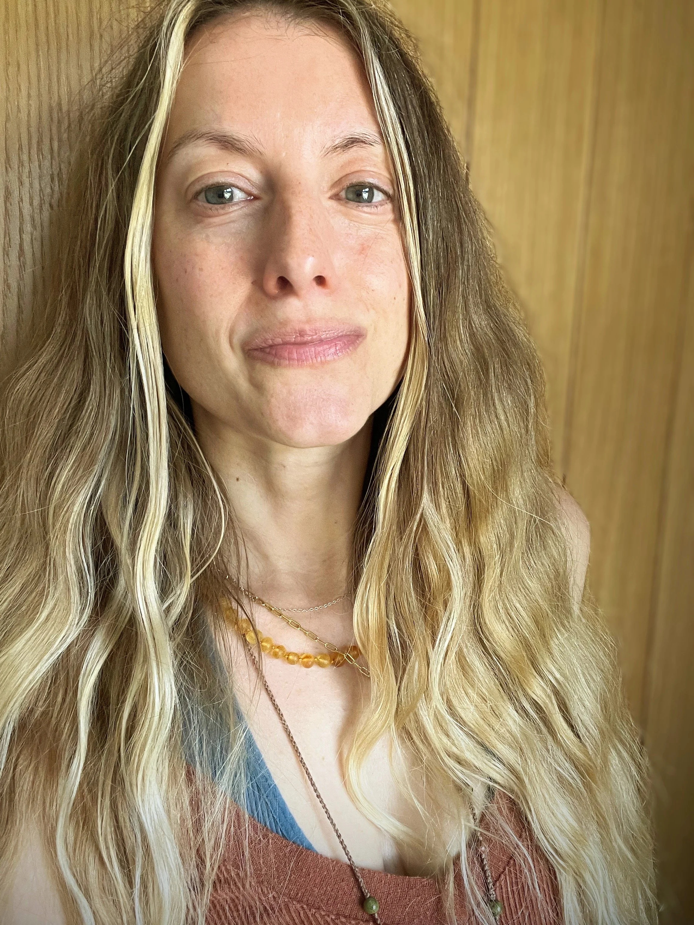 A woman with long, wavy blonde hair and light complexion, wearing a brown top and layered necklaces, standing against a wooden background.