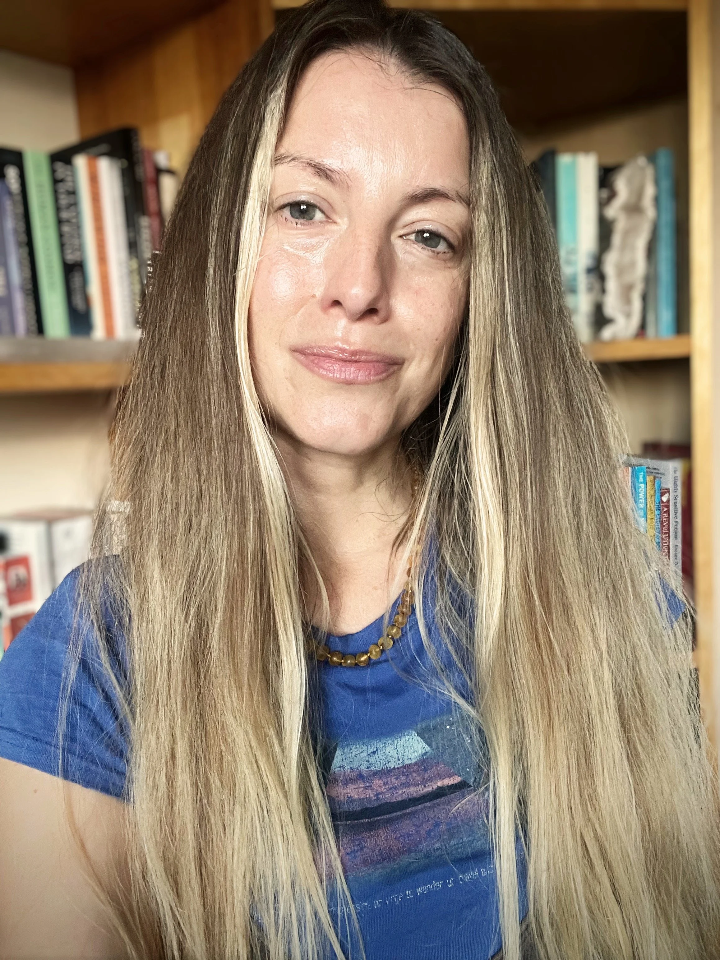 A woman with long blonde hair, wearing a blue shirt and a yellow beaded necklace, is smiling softly in front of a bookshelf filled with books.