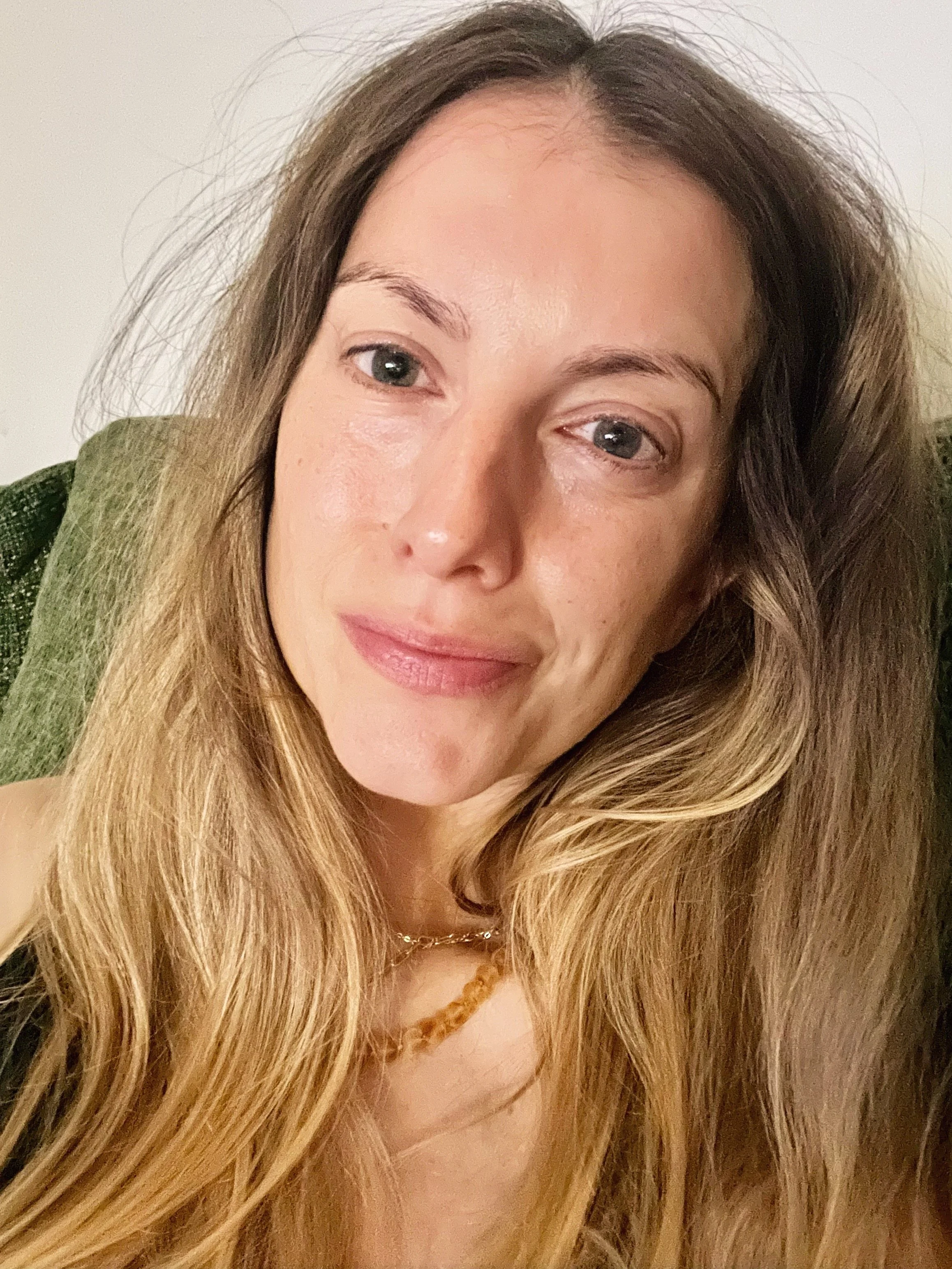 A woman with long, wavy blonde hair and blue eyes sitting on a green sofa, smiling softly at the camera.