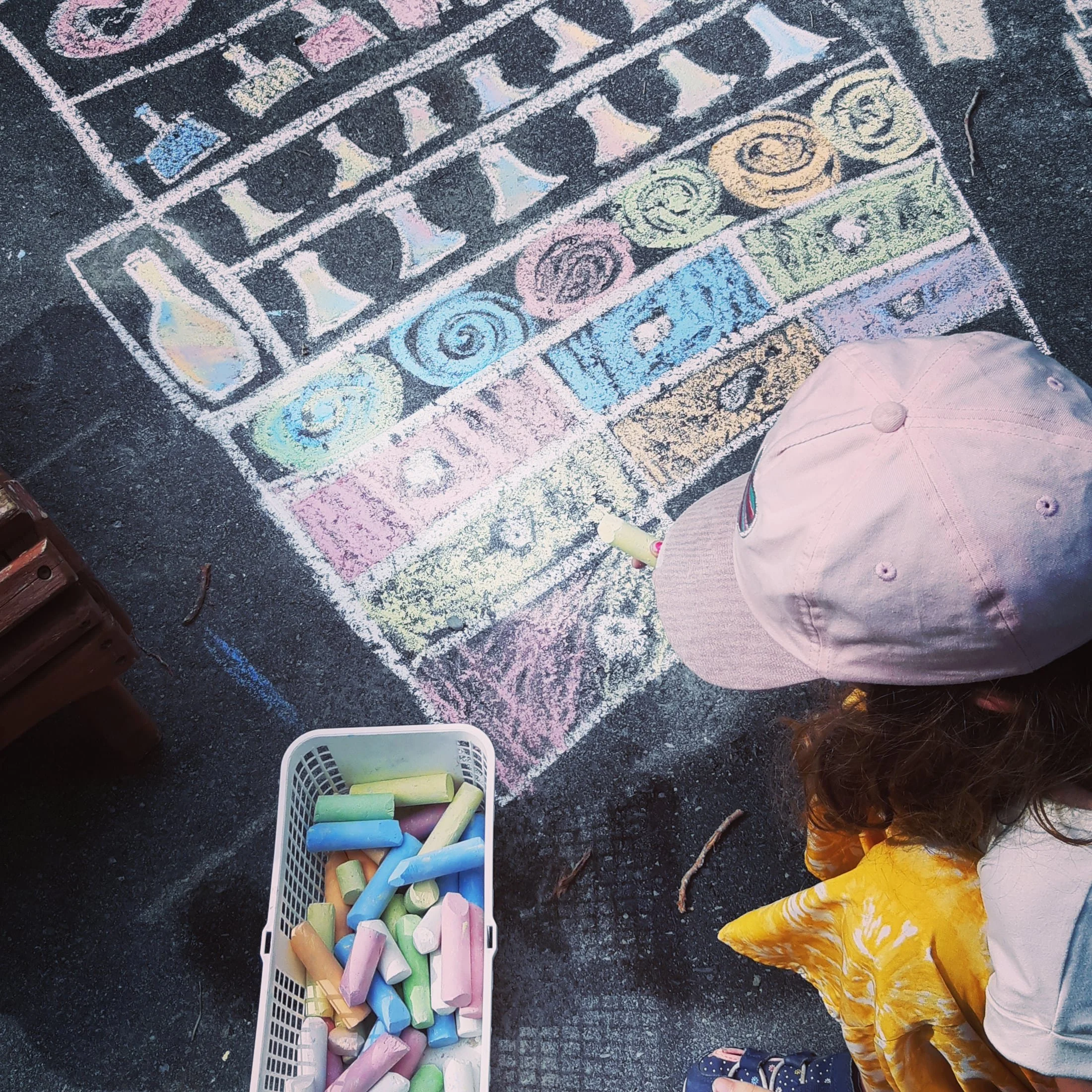Colorful chalk pastels in a white basket on a black asphalt ground, with children wearing bright clothing and sandals nearby.
