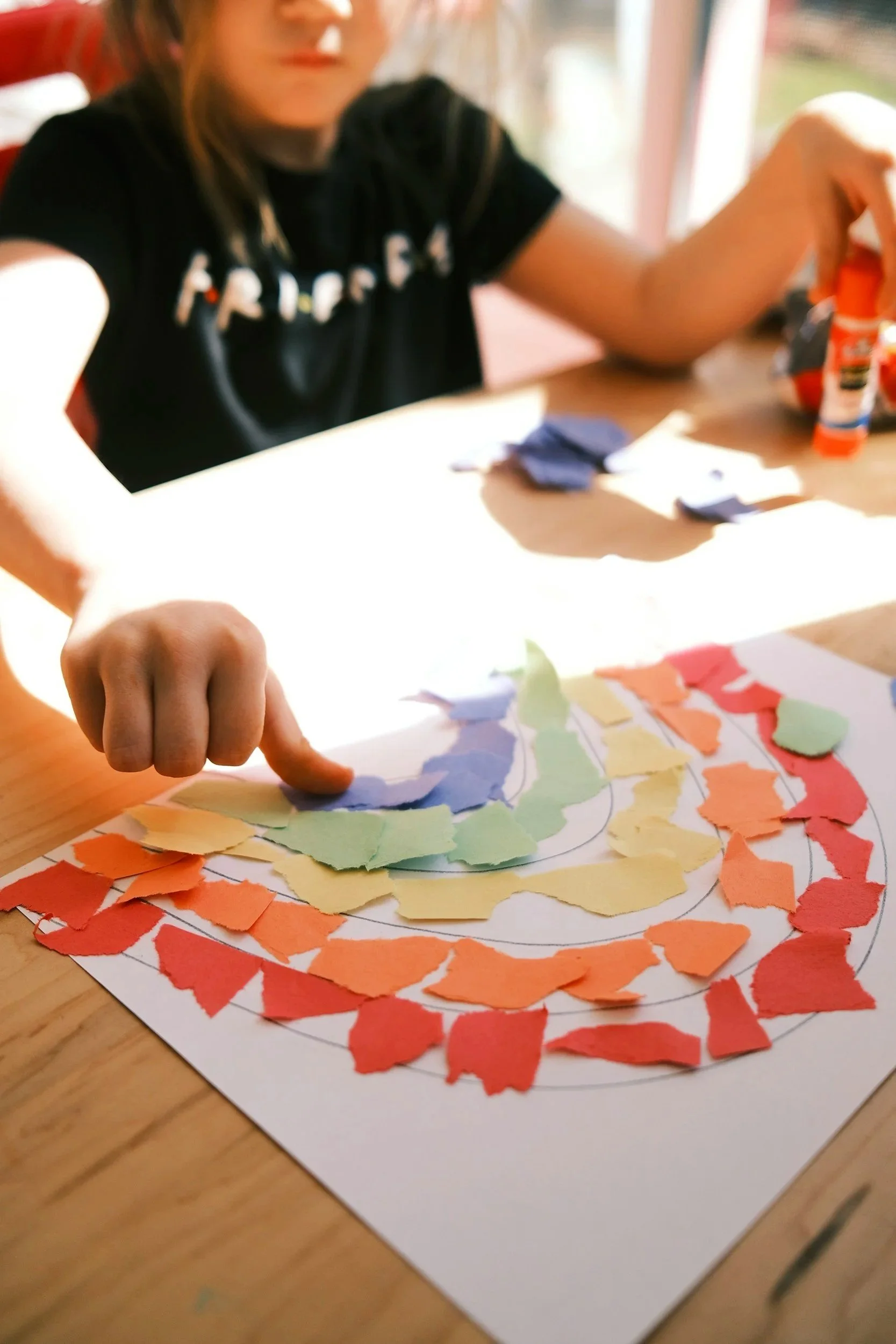 Child arranging torn paper pieces in a rainbow pattern on a sheet of paper, creating a colorful mosaic art project.