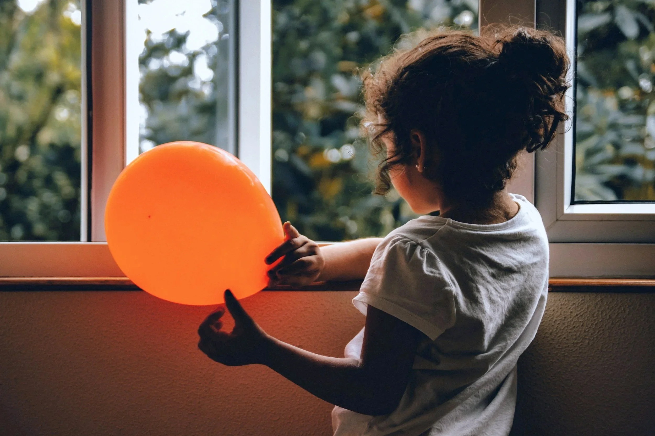 A young girl with curly hair looking out a window while holding an orange balloon.