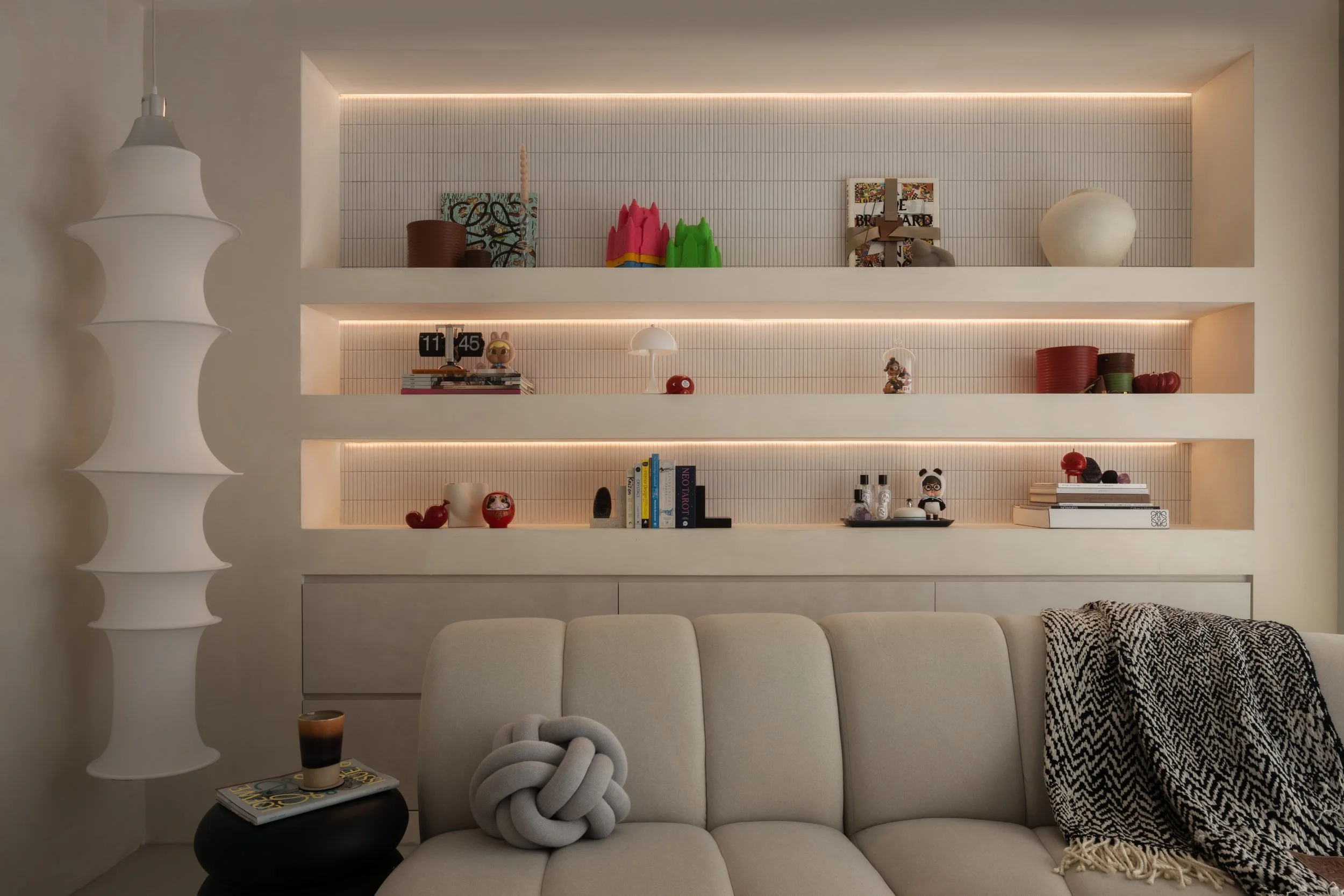 Living room wall with built-in shelves illuminated by warm lighting, decorated with books, small sculptures, and decorative objects; foreground has a beige sofa with a knot-shaped cushion and a black side table with a glass of coffee and a magazine; 
