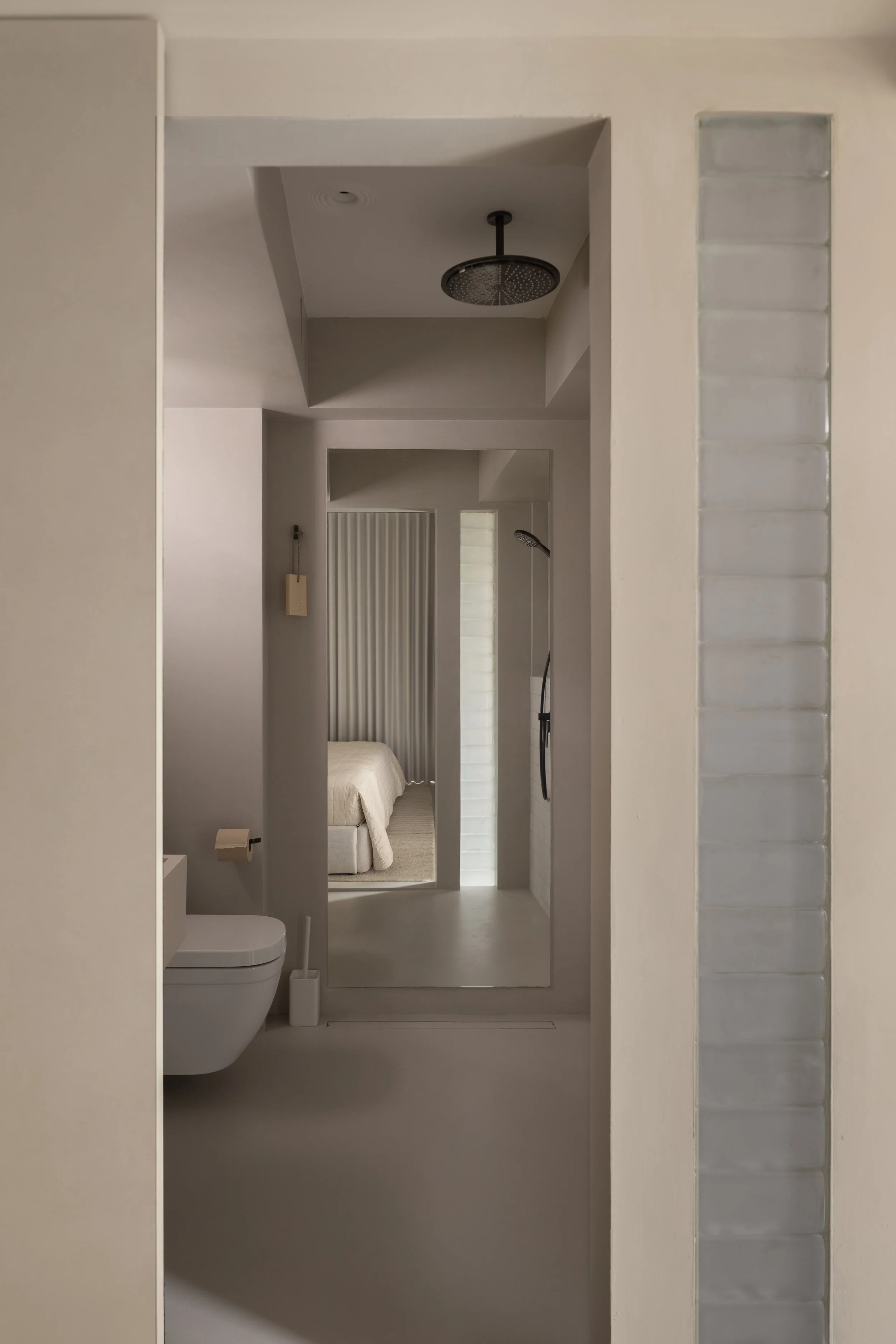 A minimalist bathroom with a white toilet, a wall-mounted shower, and a mirror reflecting a bedroom with a bed and beige bedding, with neutral tones and modern design.