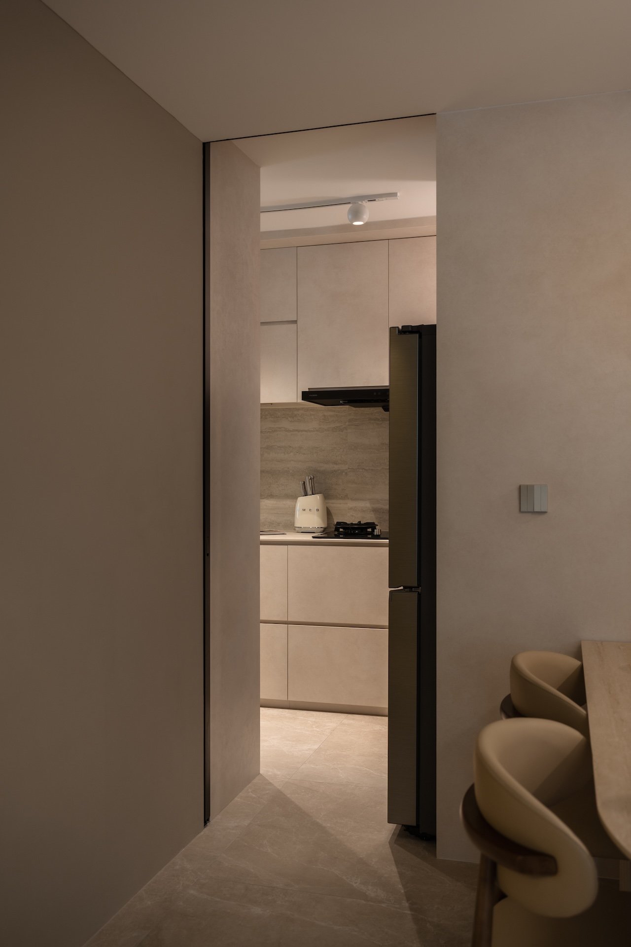 view of a modern kitchen through an open doorway, with beige cabinets, a gray backsplash, a white toaster, and a stove, with part of a dining area with beige chairs and a wooden table visible in the foreground.