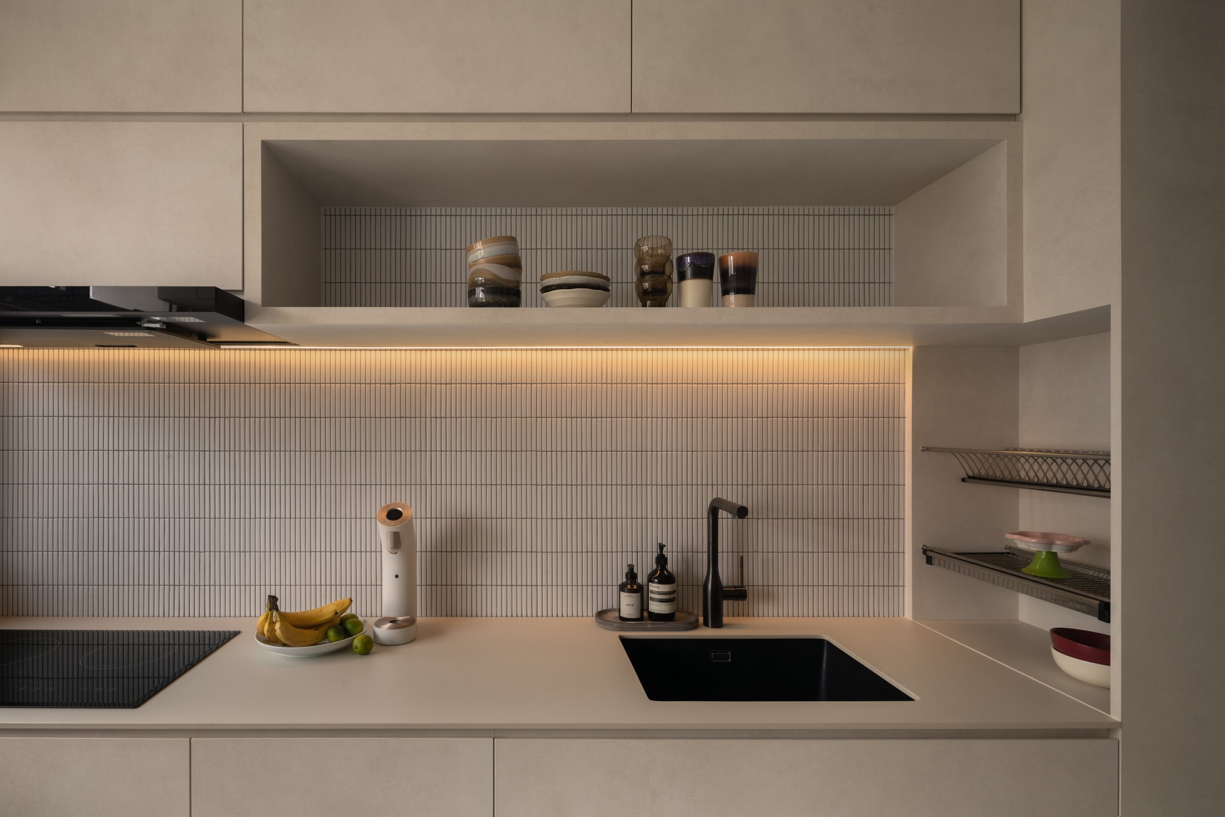 Modern kitchen with light-colored cabinets, black sink, and a countertop with bananas, limes, and a white air purifier. Open shelves with small bowls and bottles, under-cabinet lighting, and a dish rack with bowls.