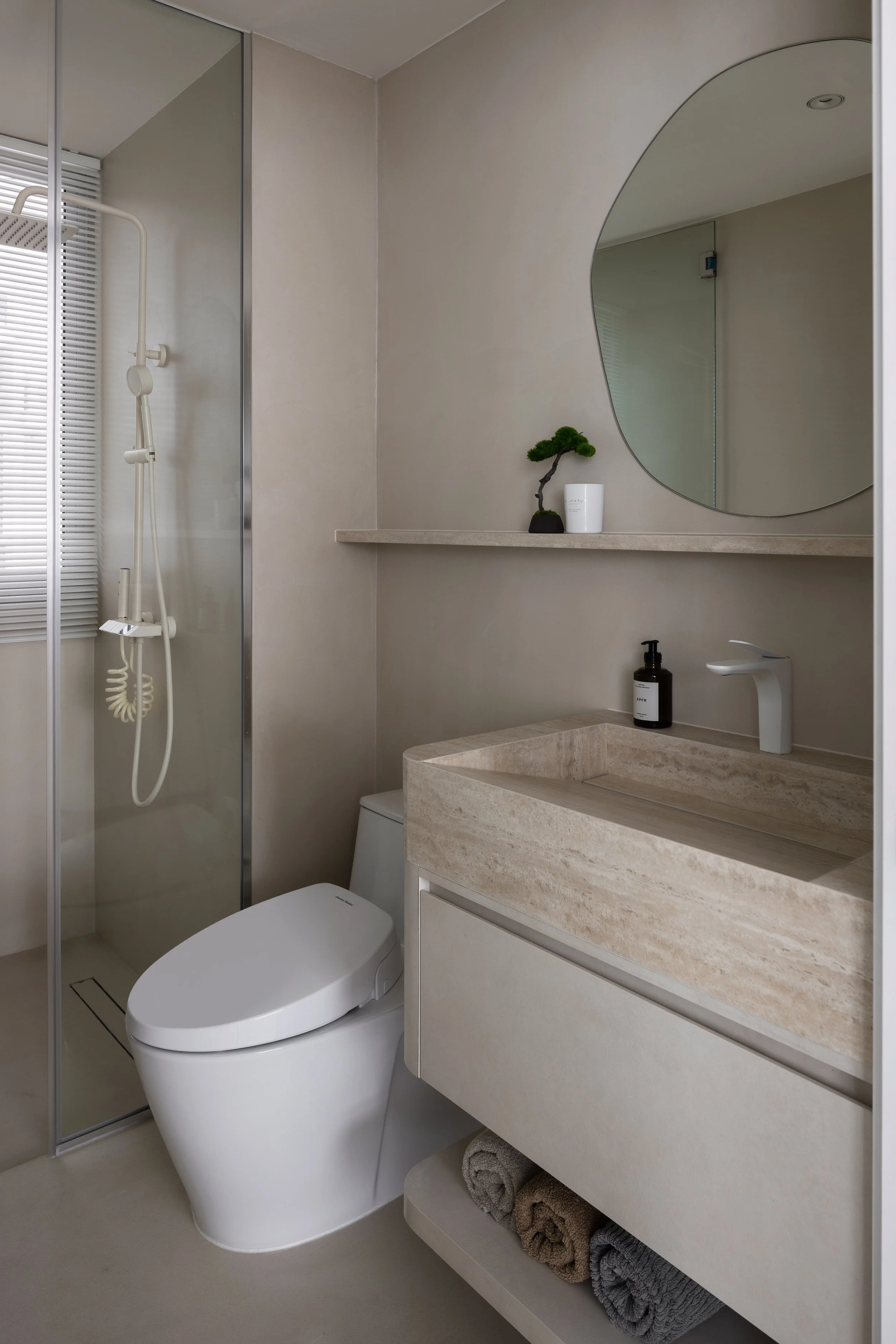 Modern bathroom with a toilet, a small sink with a black soap dispenser, a large oval mirror, a floating shelf with a small bonsai and a white container, a walk-in shower with a glass door and white fixtures, and neatly rolled towels underneath the s