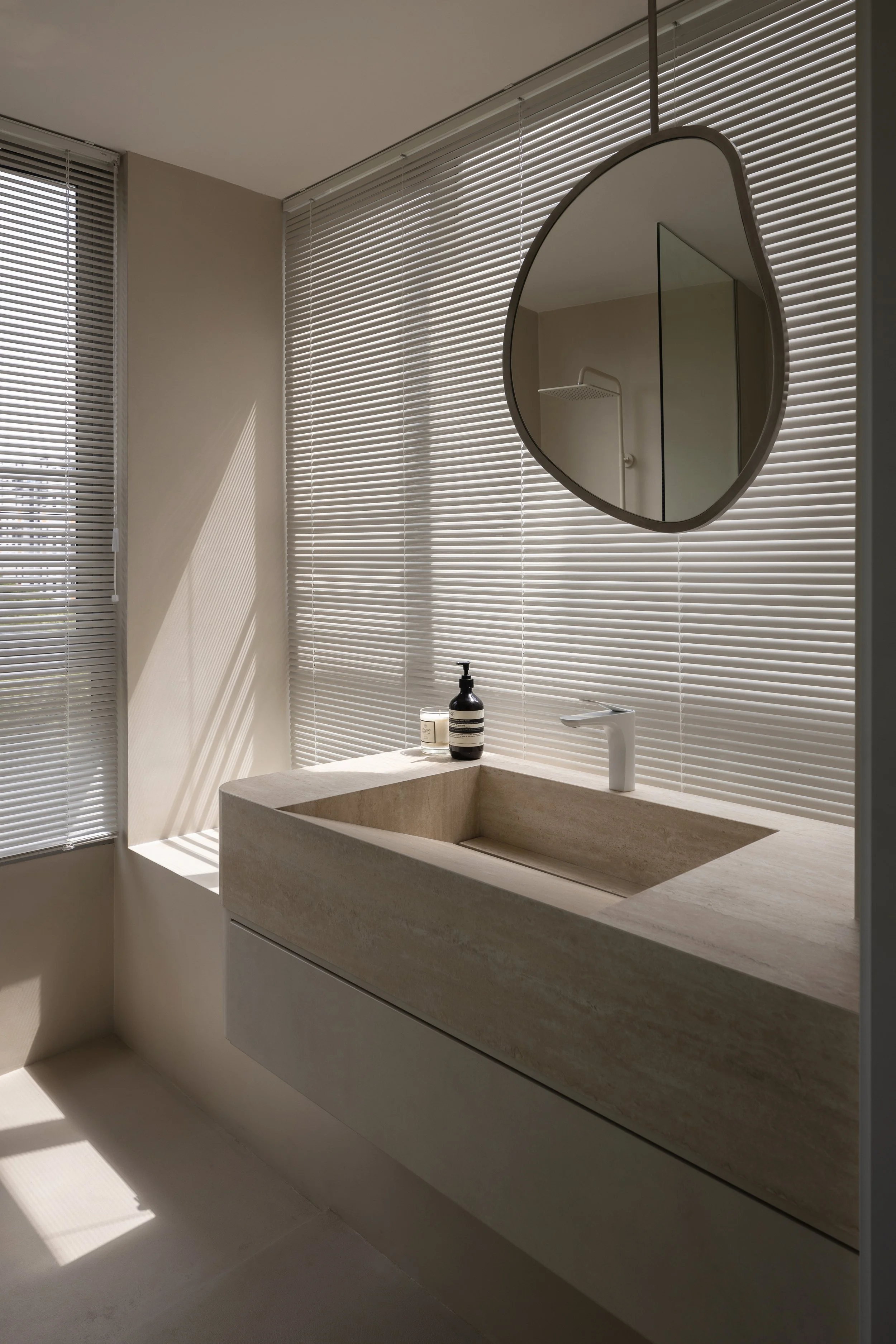 A modern bathroom with a beige stone sink, a white faucet, a round mirror, and horizontal blinds allowing sunlight to cast shadows on the wall.