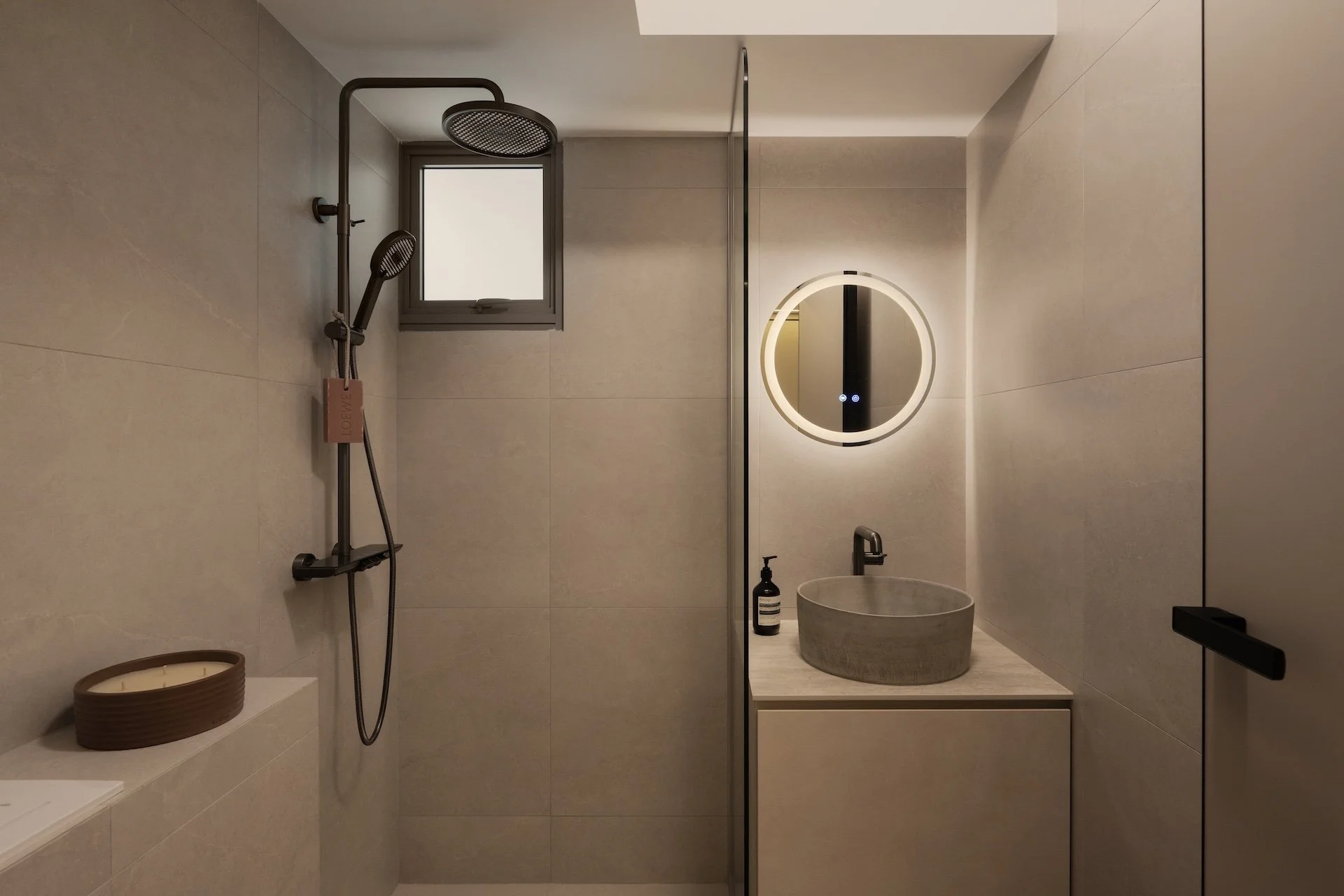 Modern minimalist bathroom with walk-in shower, round mirror with backlighting, stone sink, beige tile walls, small window, black soap dispenser, and hidden storage cabinets.
