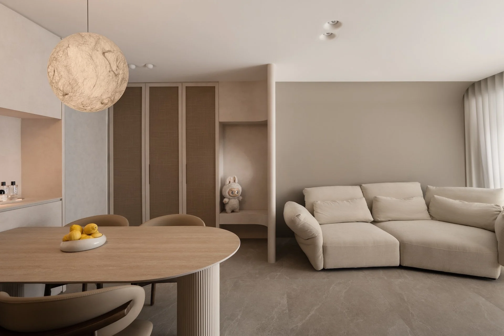 A modern living room and dining area with a round wooden dining table, beige chairs, a white sofa, and a plush bunny toy on a small shelf. The room features a large spherical pendant light and neutral-toned decor.