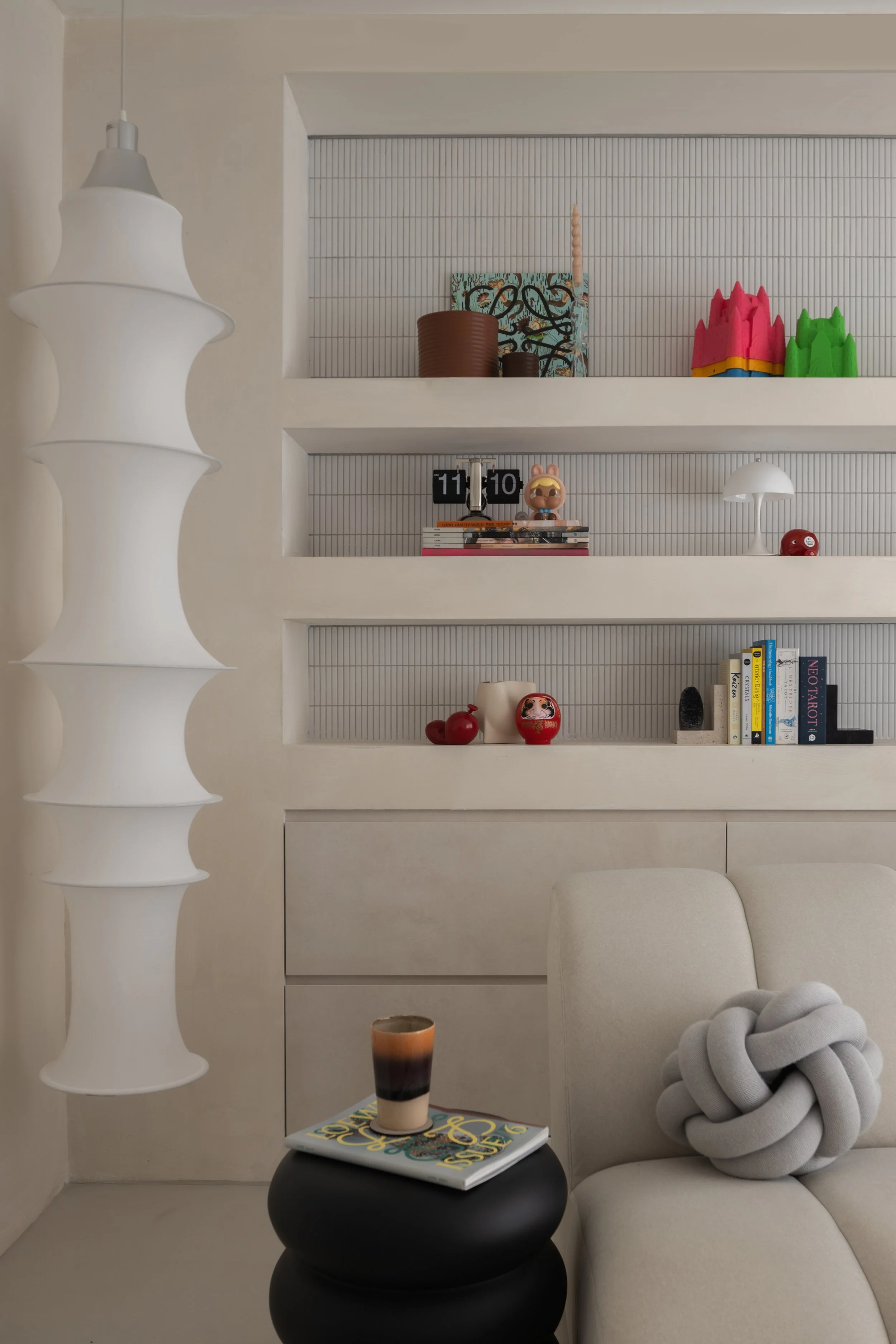 Interior living room with a white sofa, a black side table with a glass of layered beverage and magazine, and built-in white shelves with decorative items, books, and colorful ornaments.