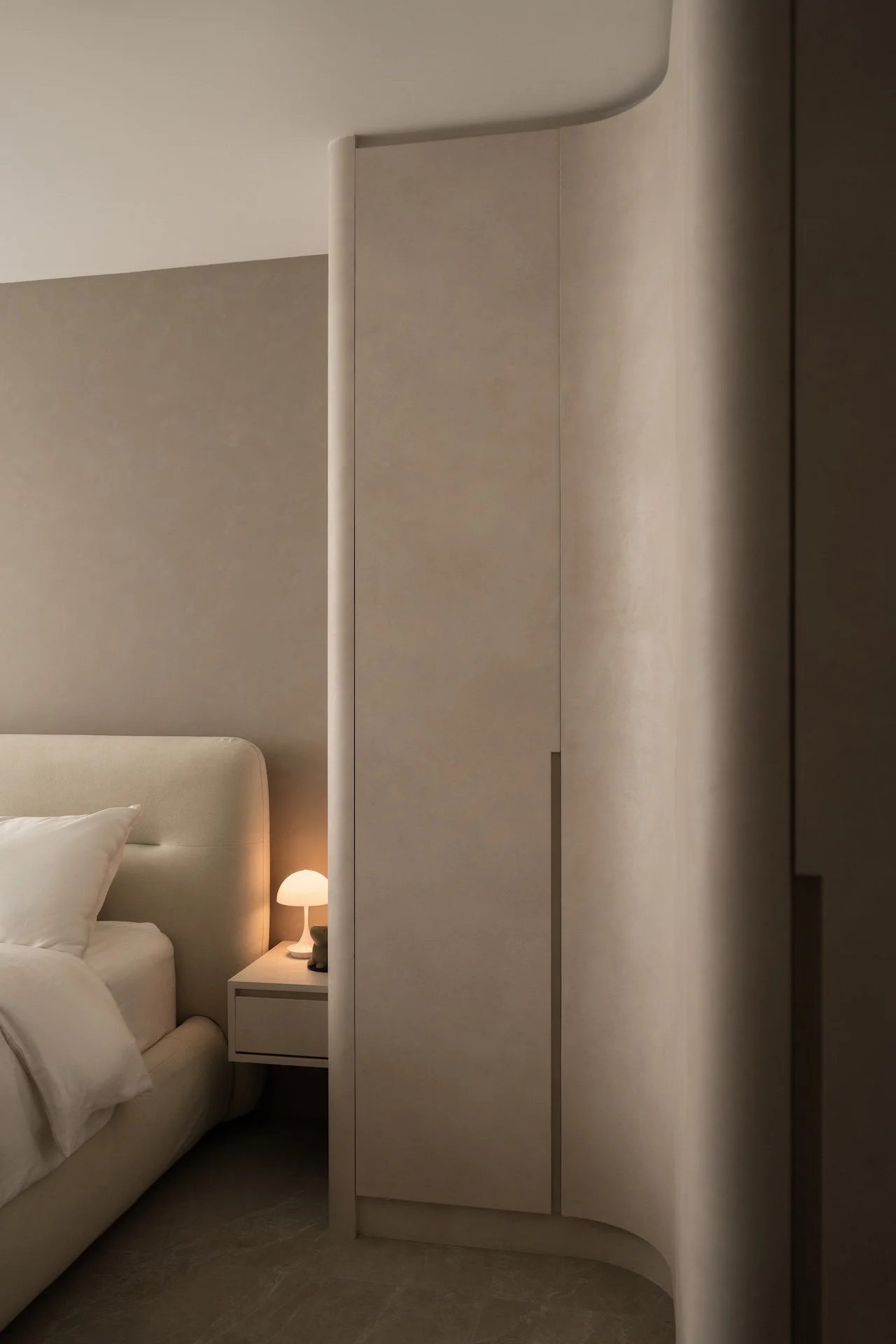 A bedroom with a beige headboard, white bedding, a white nightstand, and a small lamp. There is a curved beige room divider or wall next to the bed.