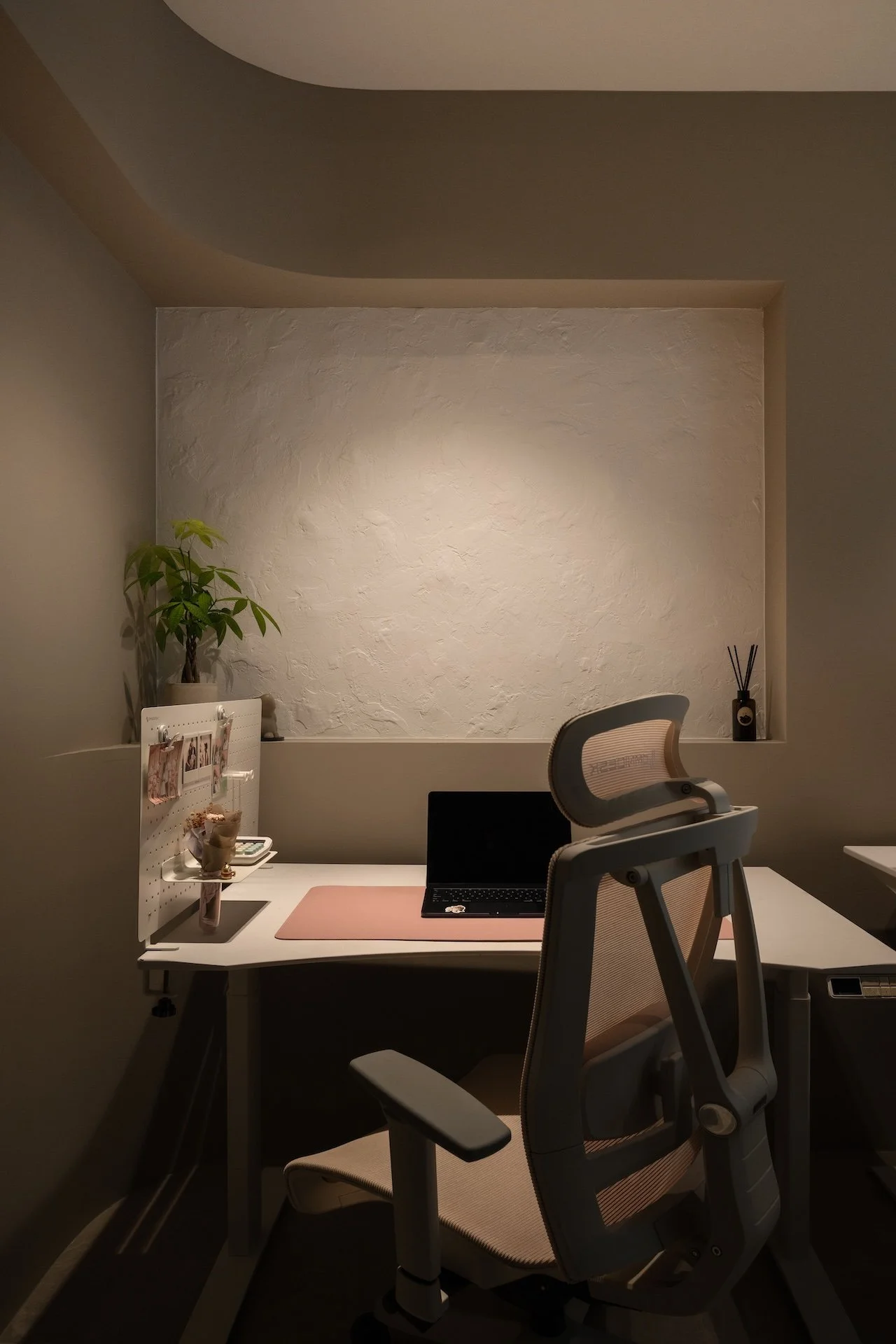 A minimalist home office with a white desk, an ergonomic office chair, a laptop, and a small decorative plant on the left. The background features a textured white wall with a recessed nook containing a small reed diffuser and a black container with 