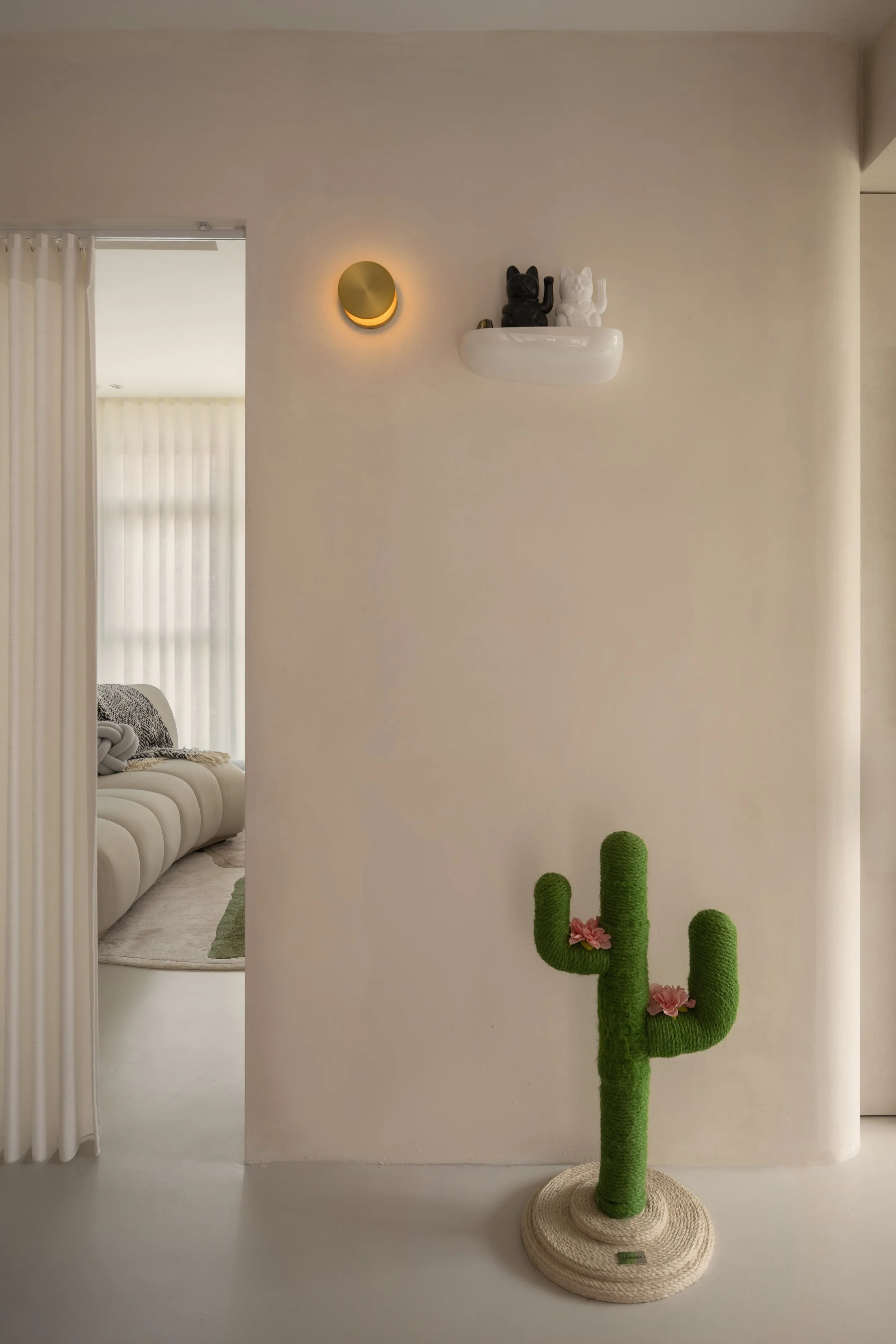 Interior scene with a white wall, a decorative cactus made of green yarn, small pink flowers, a white shelf with black and white cat figurines, a modern wall light, and a doorway with curtains leading to a living room with a cream-colored sofa and a 