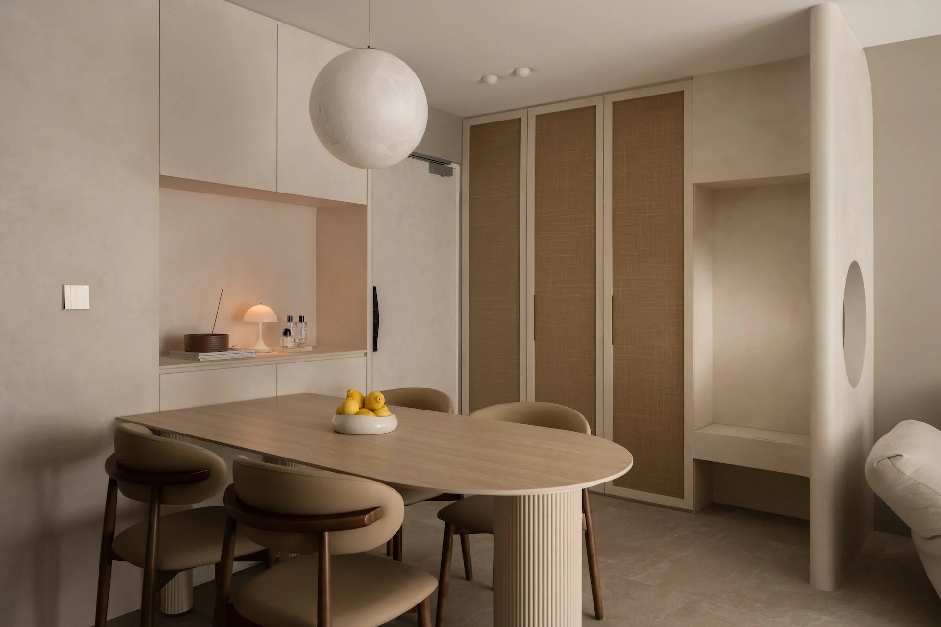 Minimalist dining room with oval wooden table, four beige chairs, a small bowl of lemons, a built-in cabinet with a lamp and decorative items, beige walls, and a large spherical hanging light.