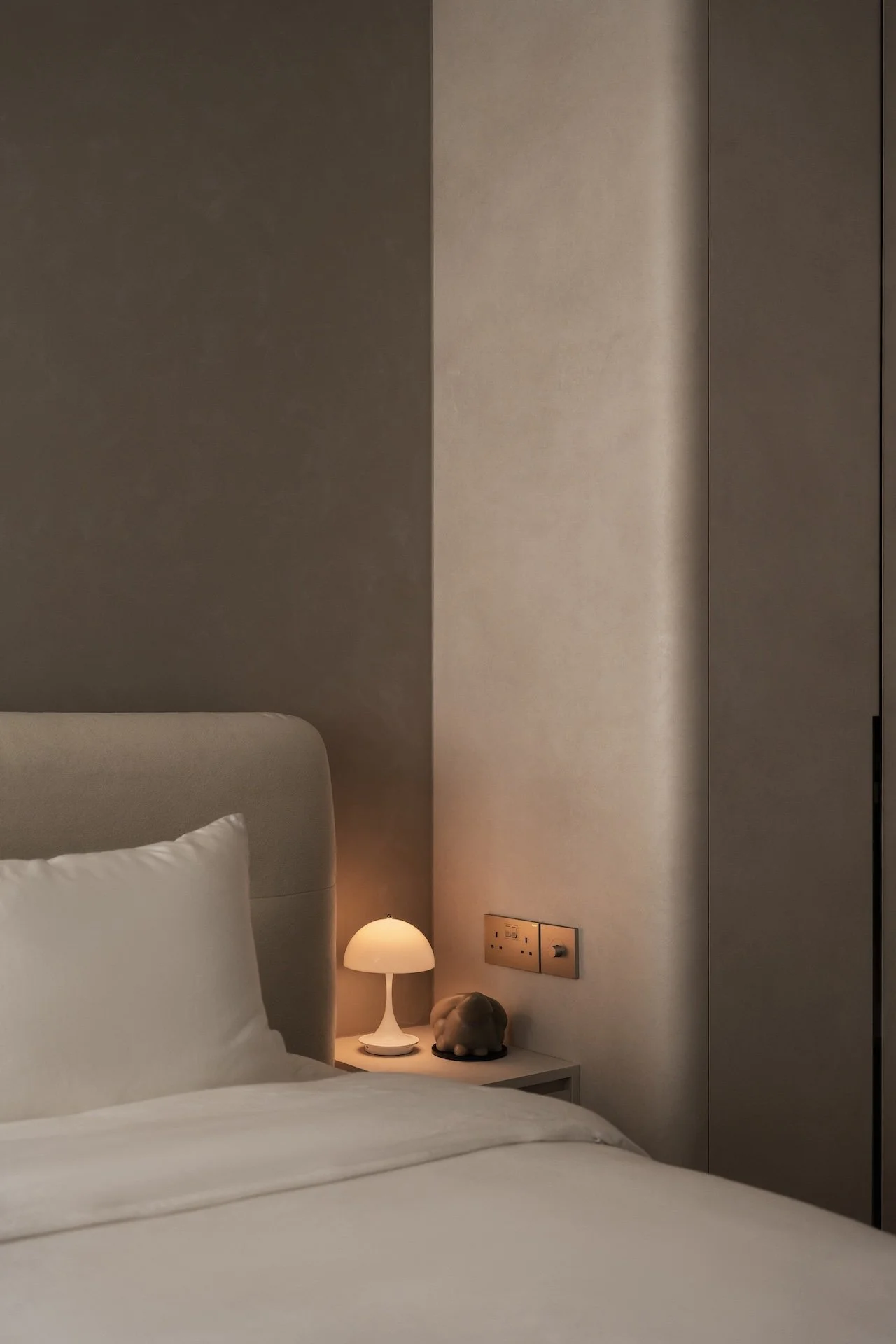 A neatly made bed with a beige headboard, white sheets, and a pillow, with a small white bedside table holding a lamp and a decorative turtle figurine, against a wall with electrical outlets and a hidden door.