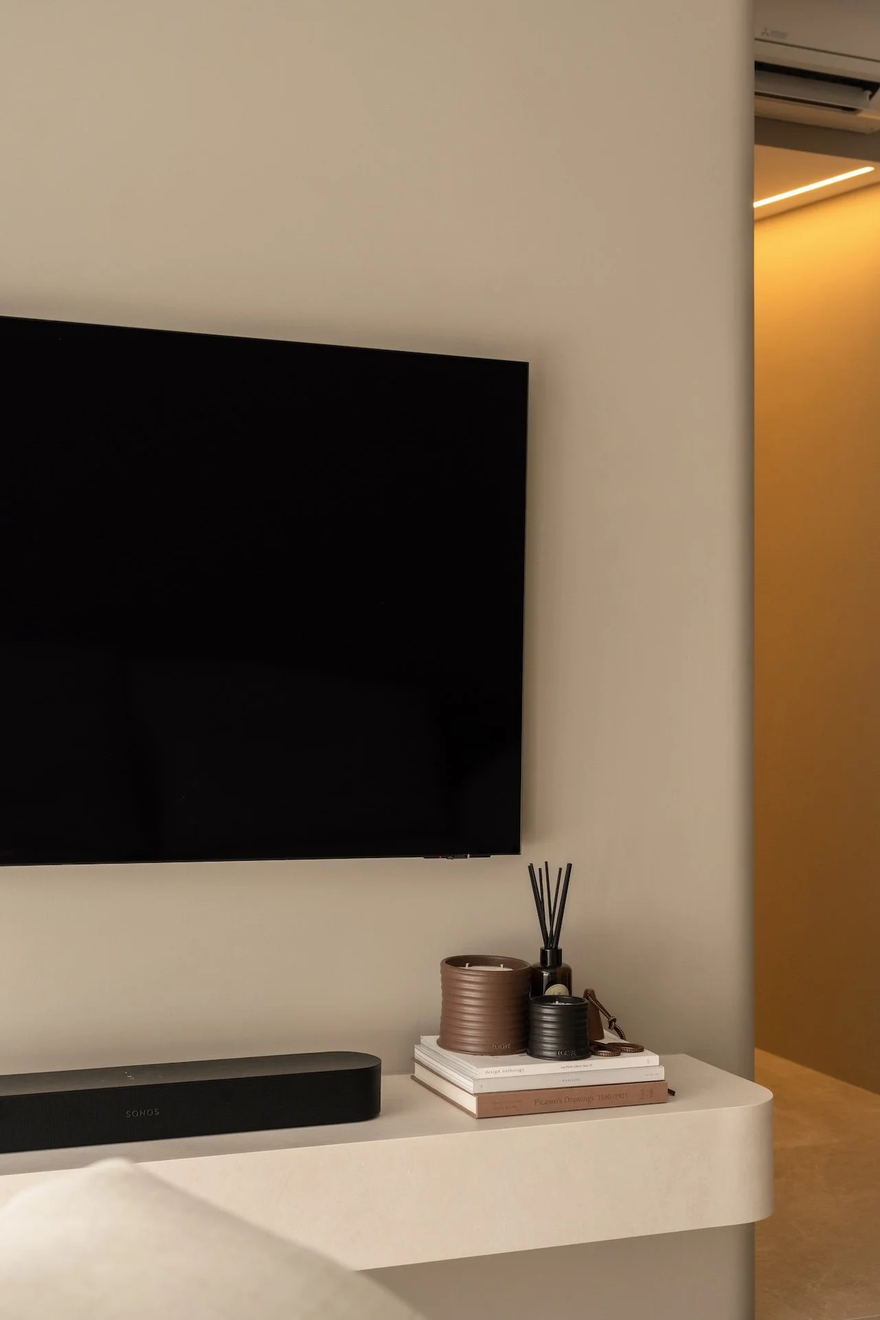 A wall-mounted flat-screen TV above a white dresser with a black speaker, decorative vases, a set of reed diffusers, and a stack of books in a modern, minimalist interior.