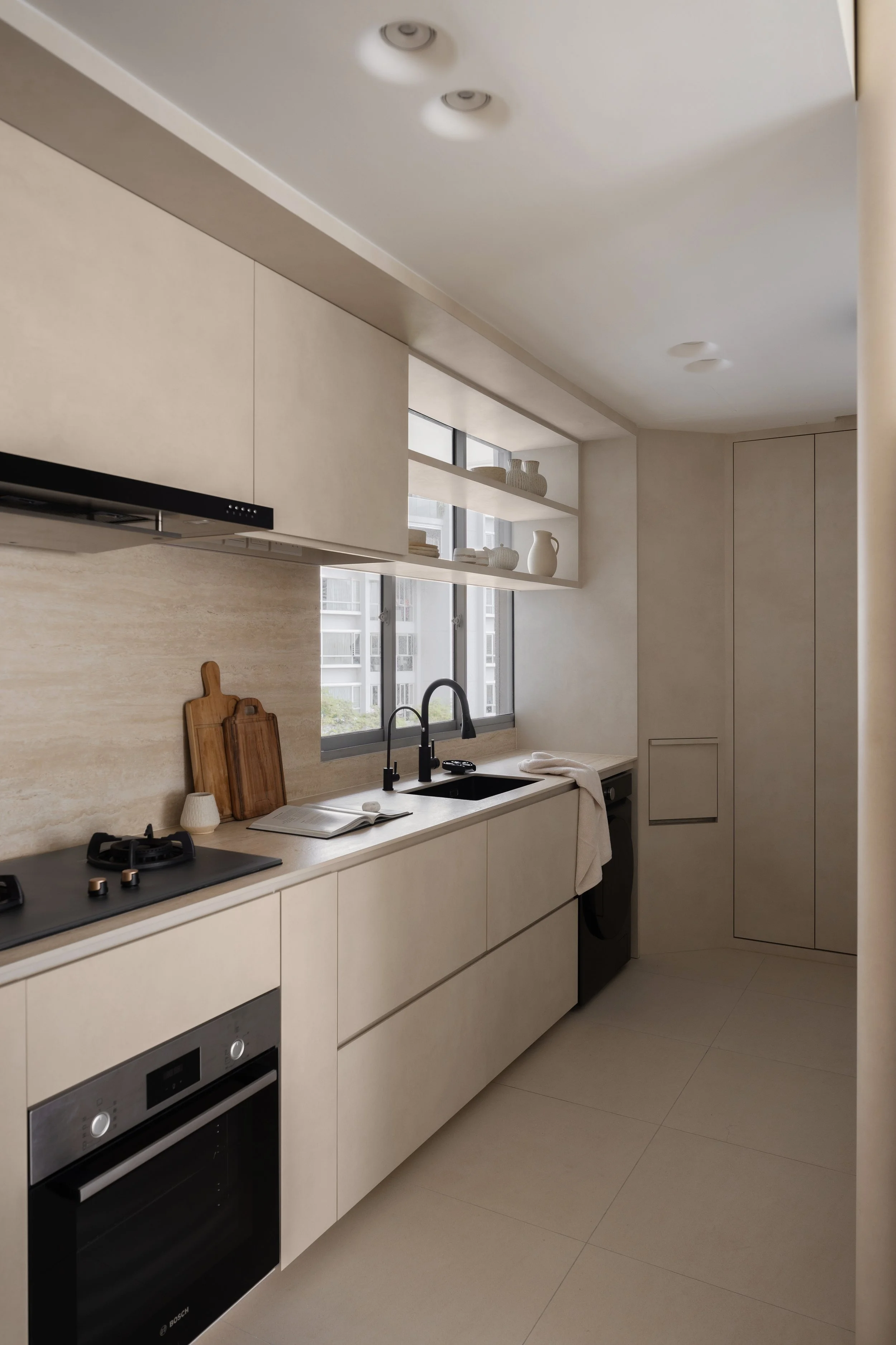 Modern kitchen with beige cabinets, black sink, and built-in oven. Open shelving with white dishes and decorative items. Large window with city view, minimal decor, neutral color palette.