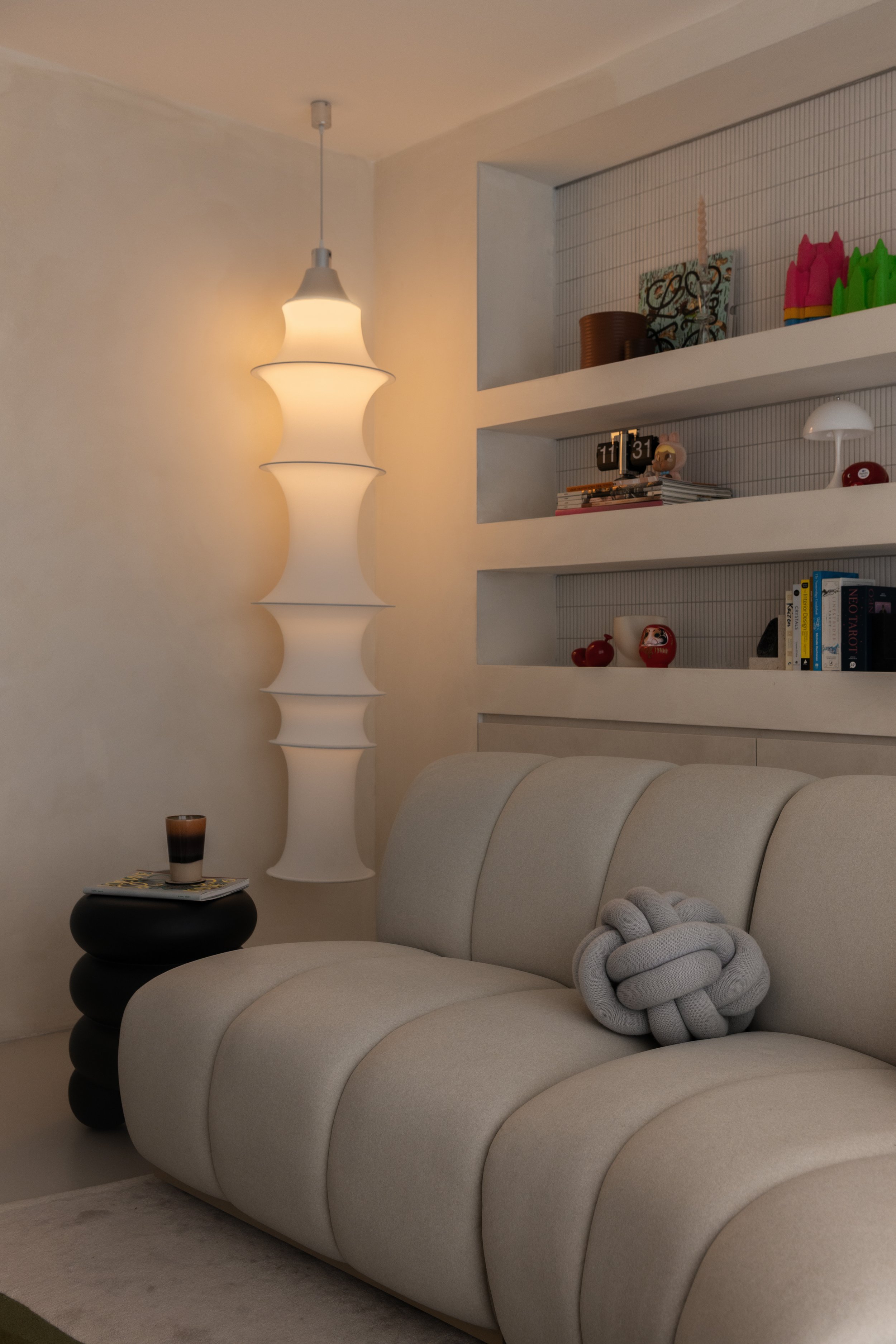 Living room with a beige sofa, black side table with a glass of drink and magazine, tall white floor lamp, built-in wall shelves with books and decorative items.
