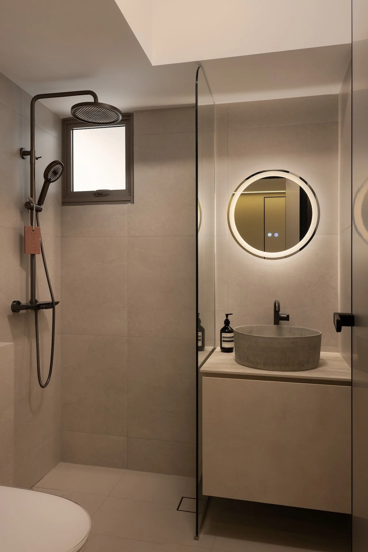 Modern bathroom with a walk-in shower featuring a rain showerhead, a small window, a round illuminated mirror, a stone sink with two black soap dispensers, and a beige cabinet.