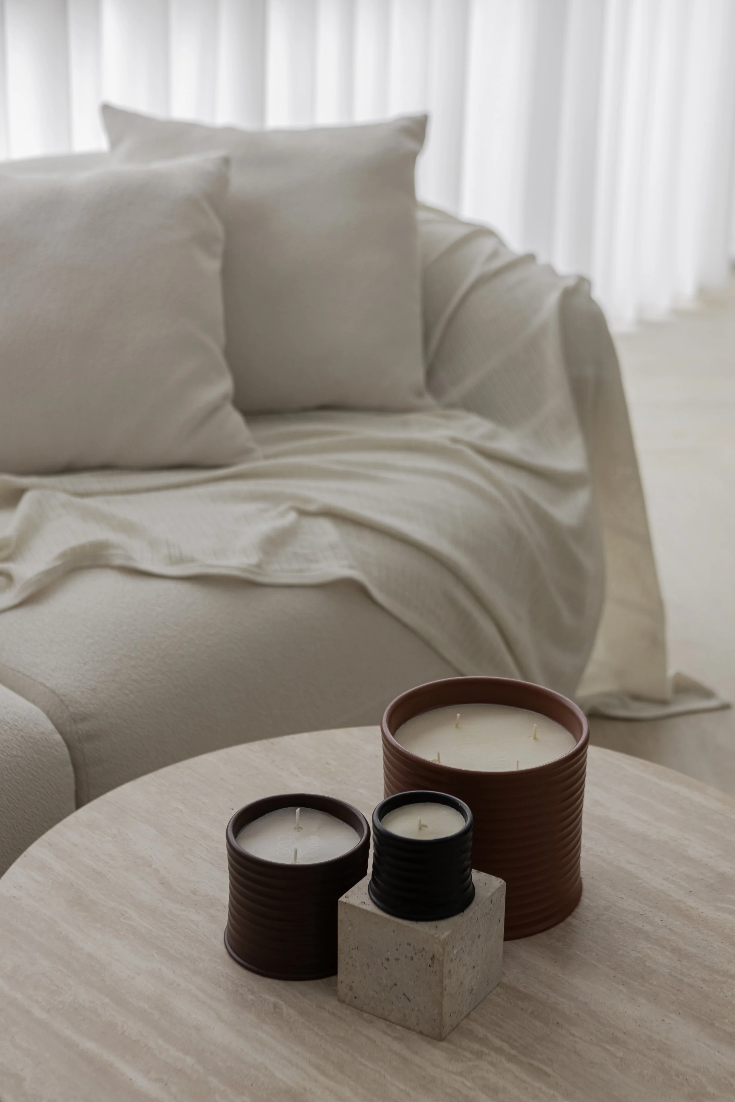 A beige sofa with white pillows and a throw blanket in a light-filled room. On a round wooden table in front of the sofa, three candles in different sized and colored holders, including brown, black, and a concrete square base, are displayed.