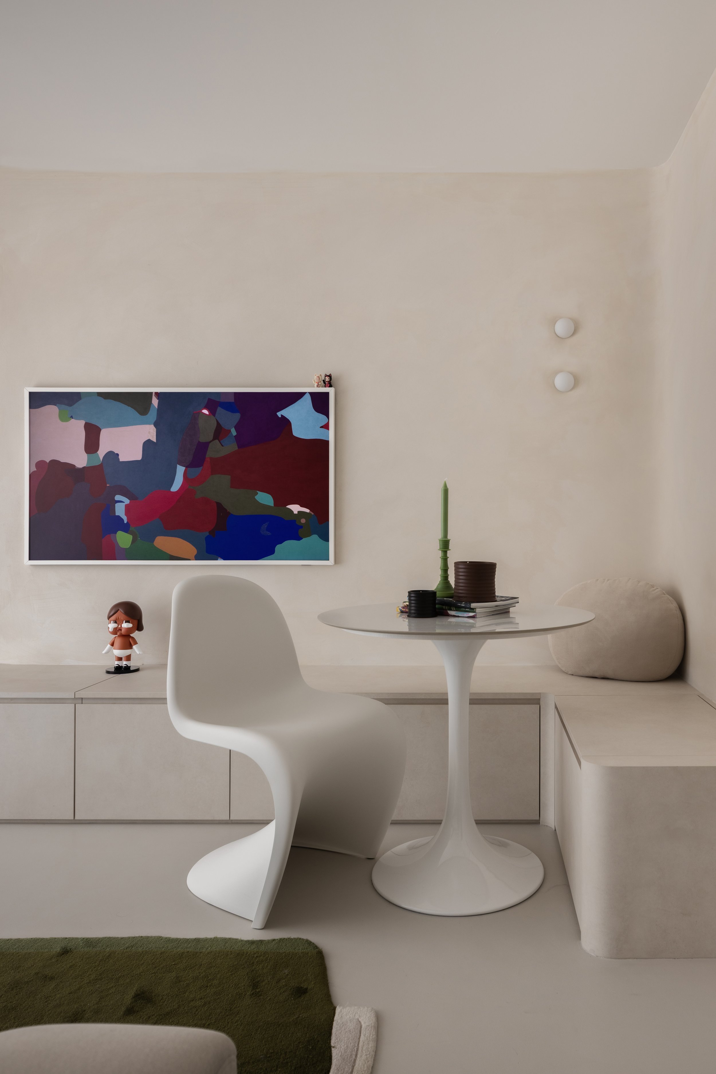 Minimalist interior with a white modern chair, round white table with a green candlestick and books, wall-mounted abstract art, and neutral decor including a small figure on the cabinet.