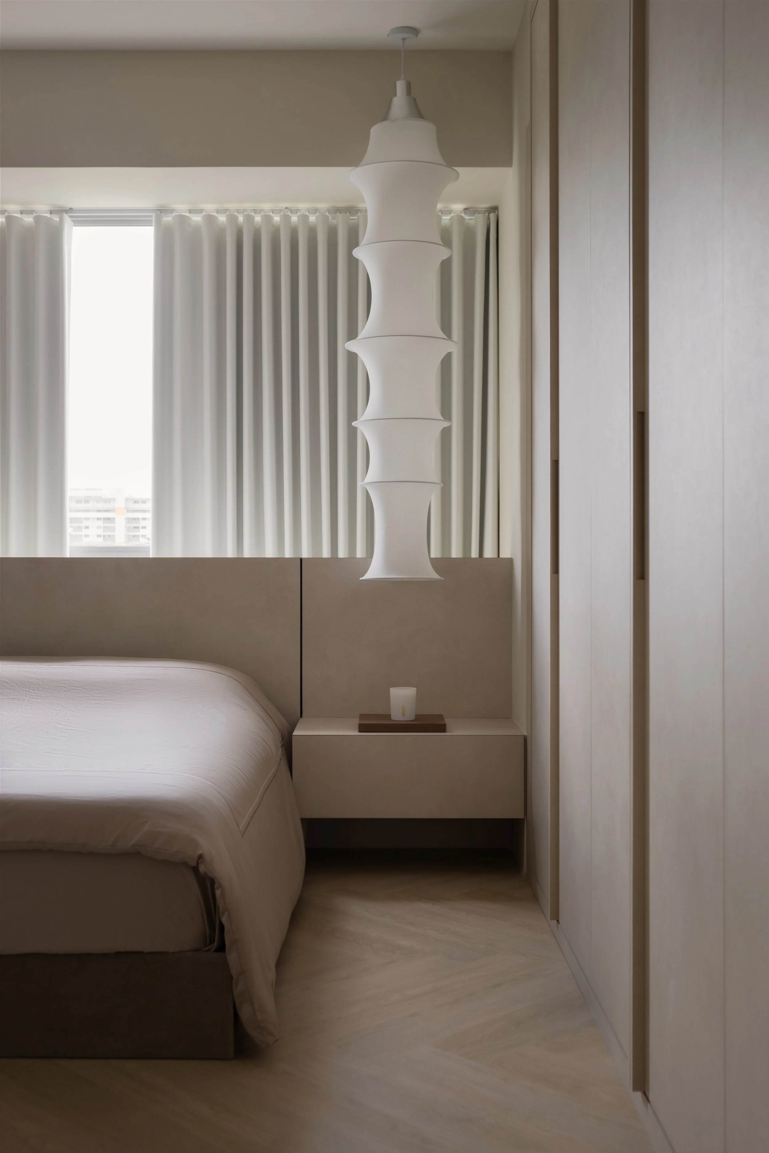Minimalist bedroom with beige bed, beige headboard, beige walls, white curtains, and a modern white hanging pendant light.