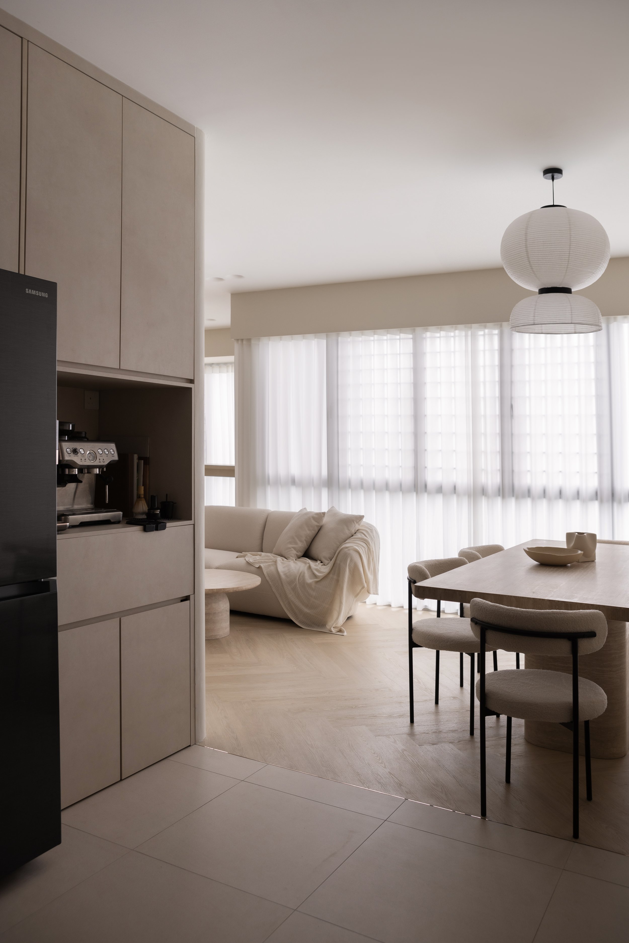 Modern living room with beige sofa, light wood dining table, and chairs, large windows with sheer white curtains, and a minimalist ceiling light fixture, with part of a kitchen visible on the left.