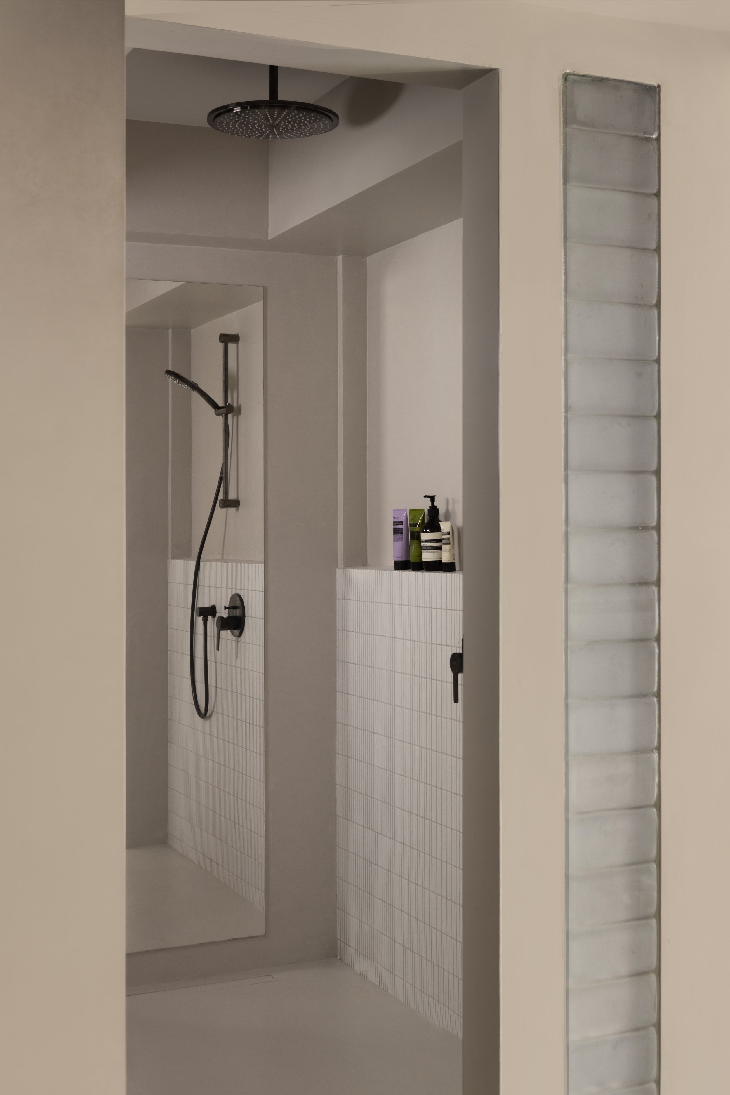 A modern walk-in shower with a black rainfall showerhead and a handheld shower, framed by a wall with white tiles and a small ledge holding toiletries.
