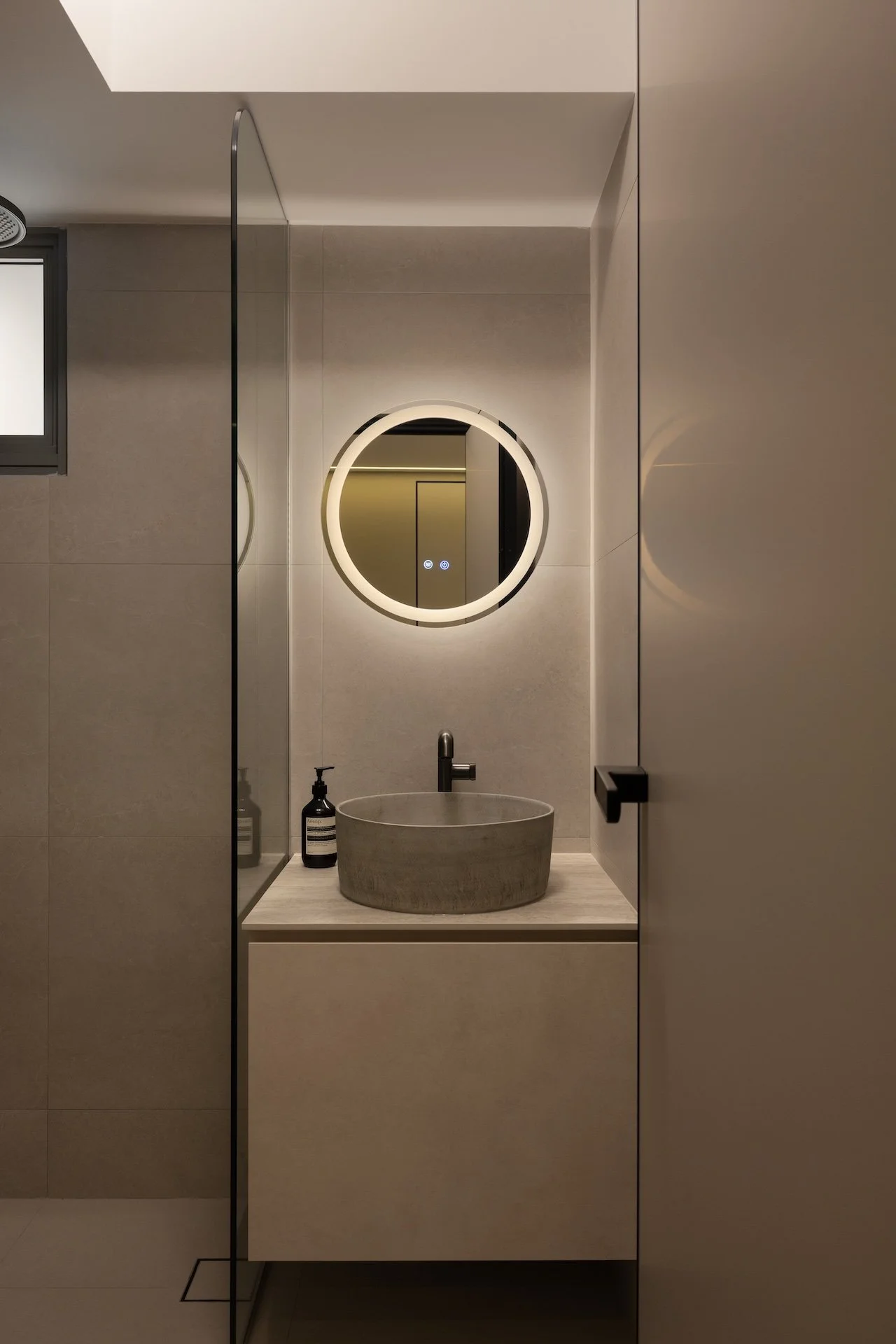 Modern bathroom with a circular mirror with LED lighting, gray vessel sink, black faucet, and a bottle of soap on the countertop, seen through a partially open door.