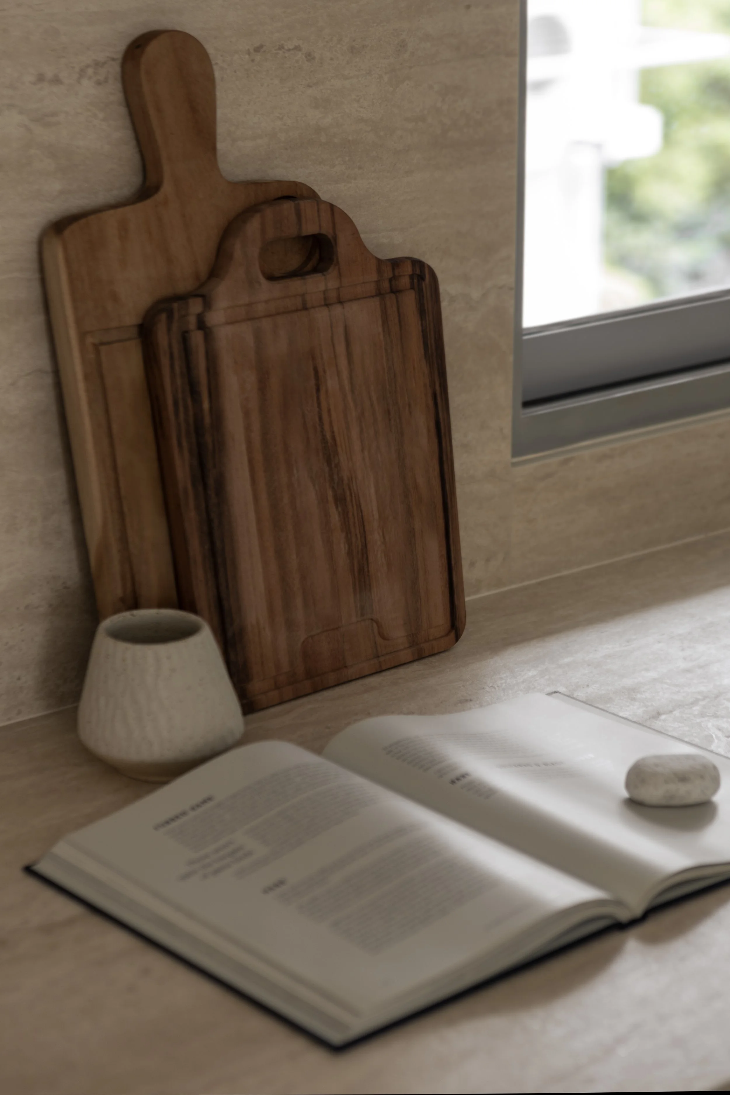 Open book on kitchen countertop with a stone, a small vase, and wooden cutting boards near window.