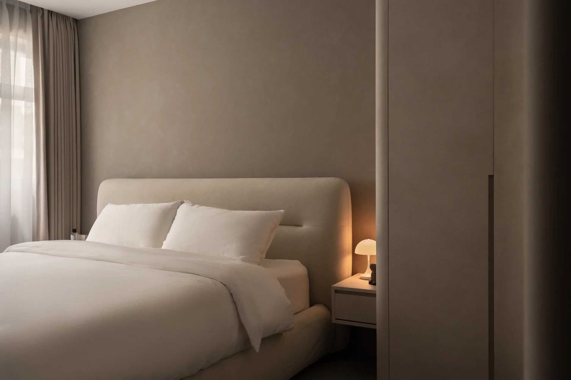 A neatly made bed with white bedding and pillows, beside a nightstand with a small lamp, in a minimalist bedroom with beige walls and curtains.