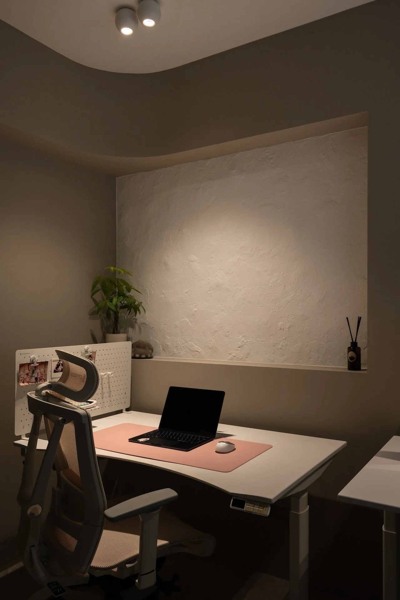 A minimalist home office with a white desk, a black laptop, a white mouse on a pink desk mat, an ergonomic chair, a small plant, a photo board, and a diffuser, with neutral-colored walls and a textured white wall feature.