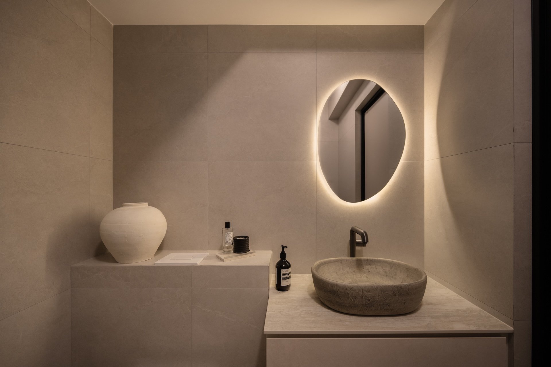 Minimalist bathroom with a circular backlit mirror, a stone vessel sink, a black faucet, a large white ceramic vase, and black soap and lotion bottles on a light-colored countertop.