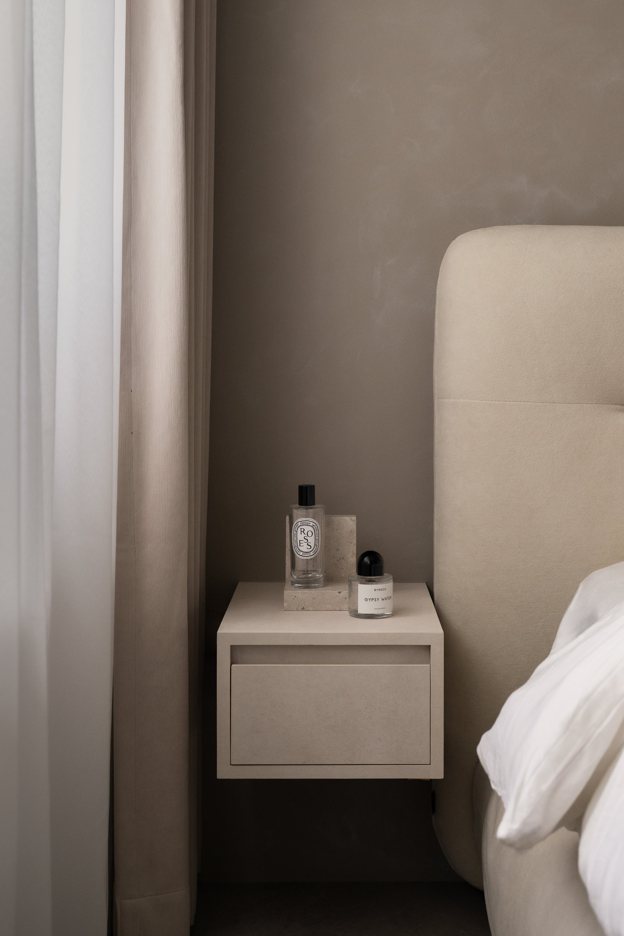 A beige nightstand next to a bed with white sheets and pillow, holding bottles of skincare or perfume; beige curtains and a gray wall in the background.