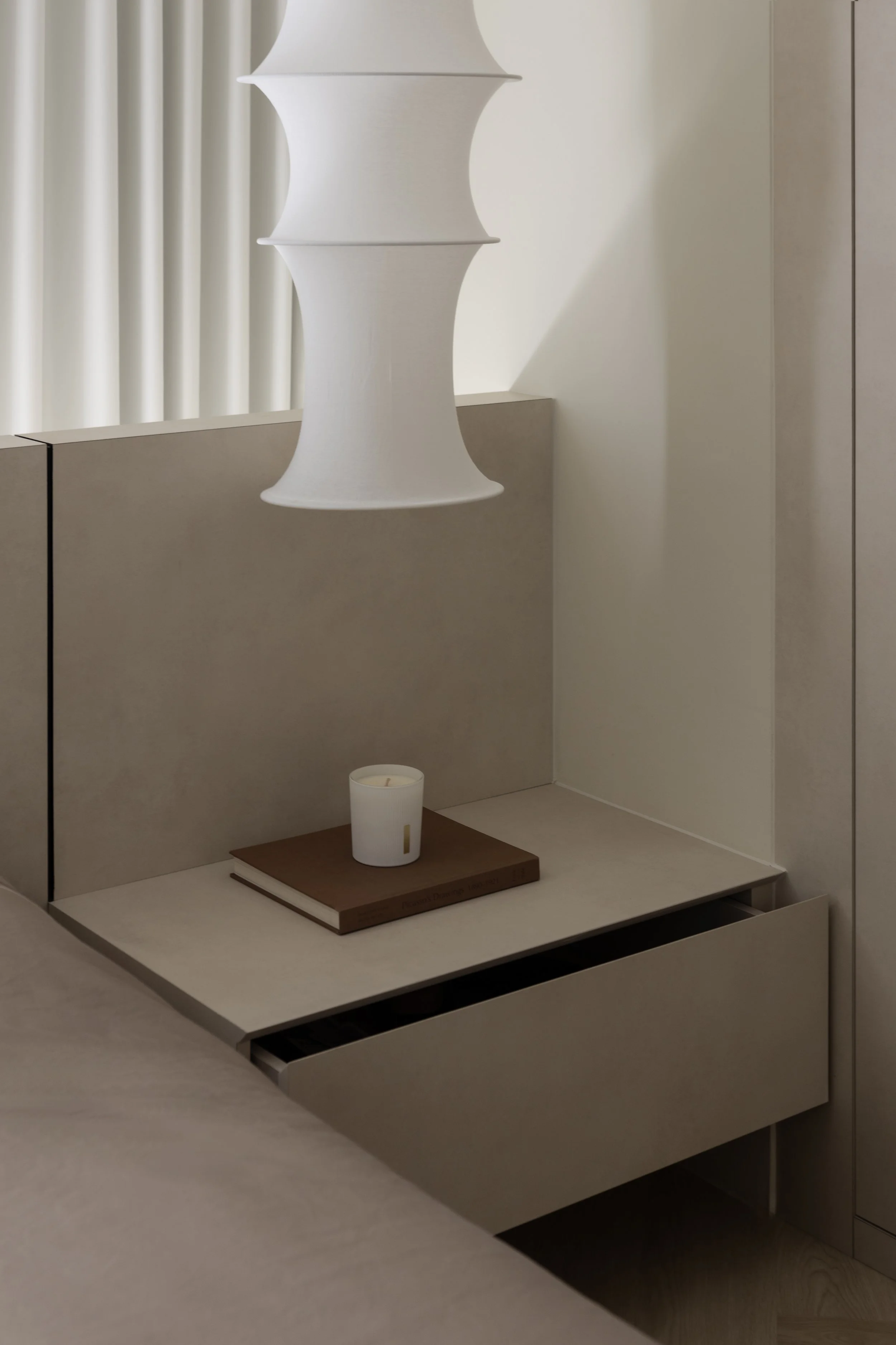 Minimalist bedroom nightstand with a white lamp, candle, and a book in a neutral-colored room.