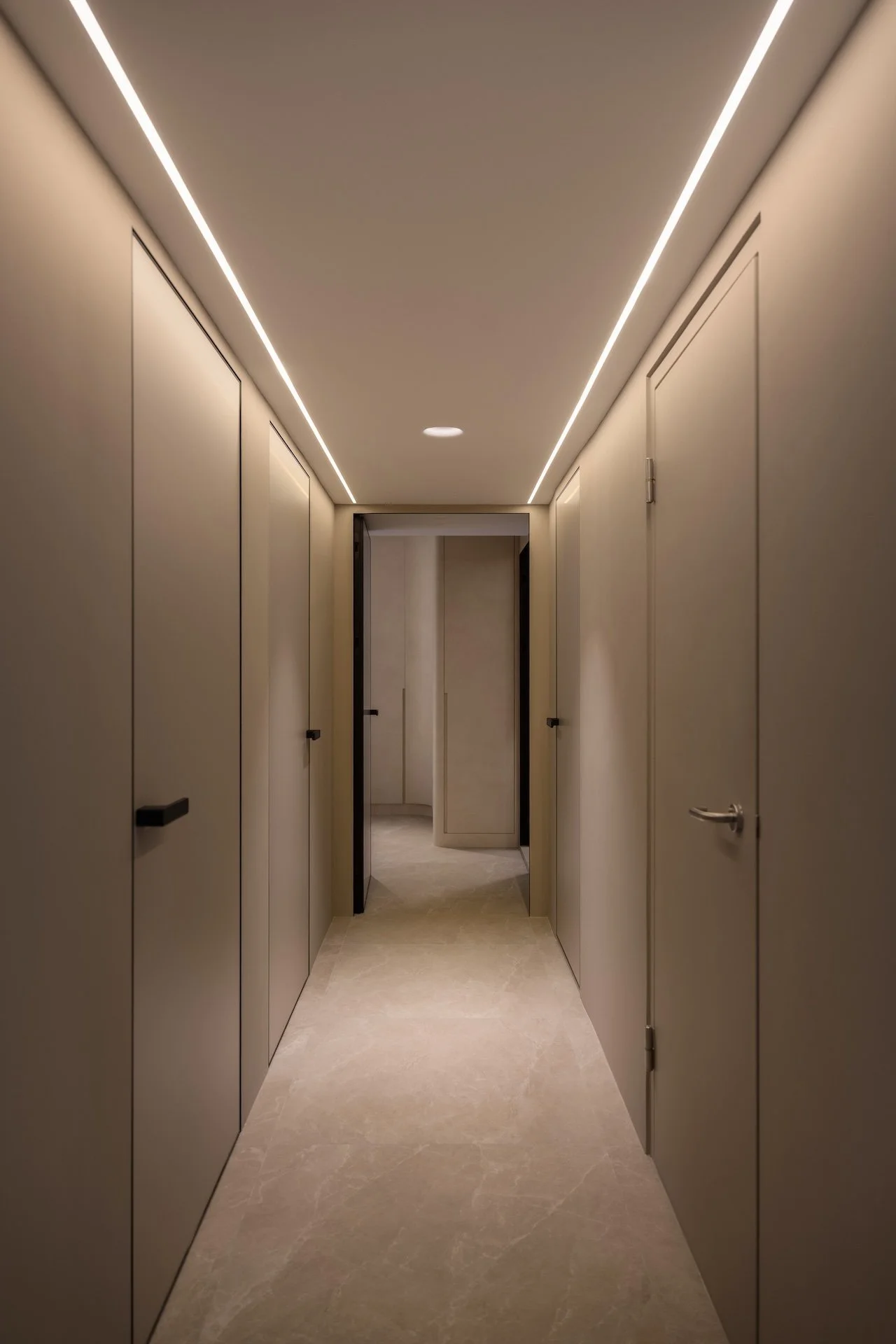 A narrow, modern hallway with beige walls, LED strip lights on the ceiling, and several closed doors on both sides, leading to a slightly open door at the end.