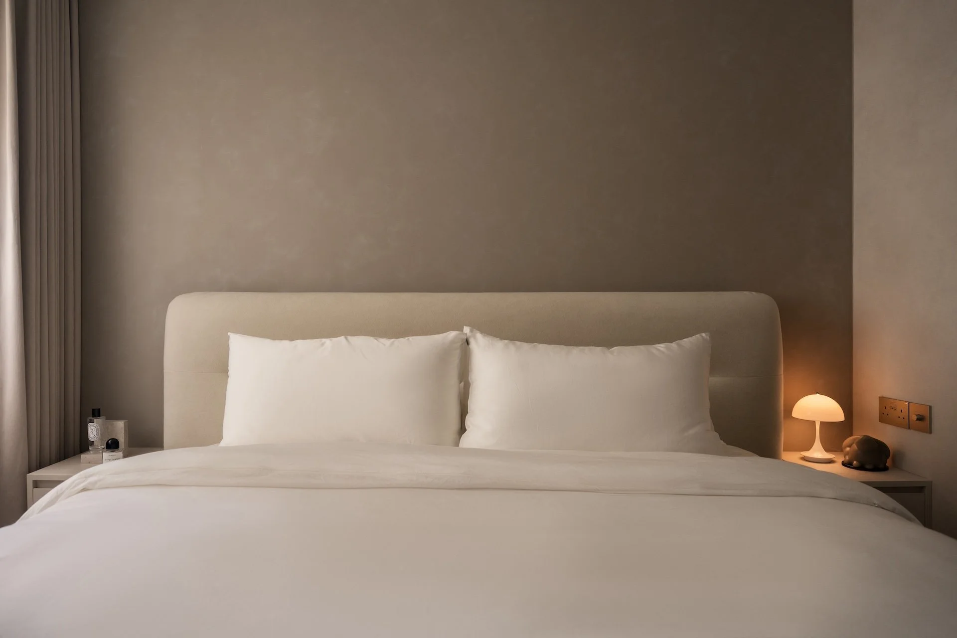 A neatly made bed with white pillows and bedding, with a small nightstand on each side. The right nightstand has a lamp, a decorative object, and electrical outlets. The beds are against a plain wall, and there is a window with curtains partially vis