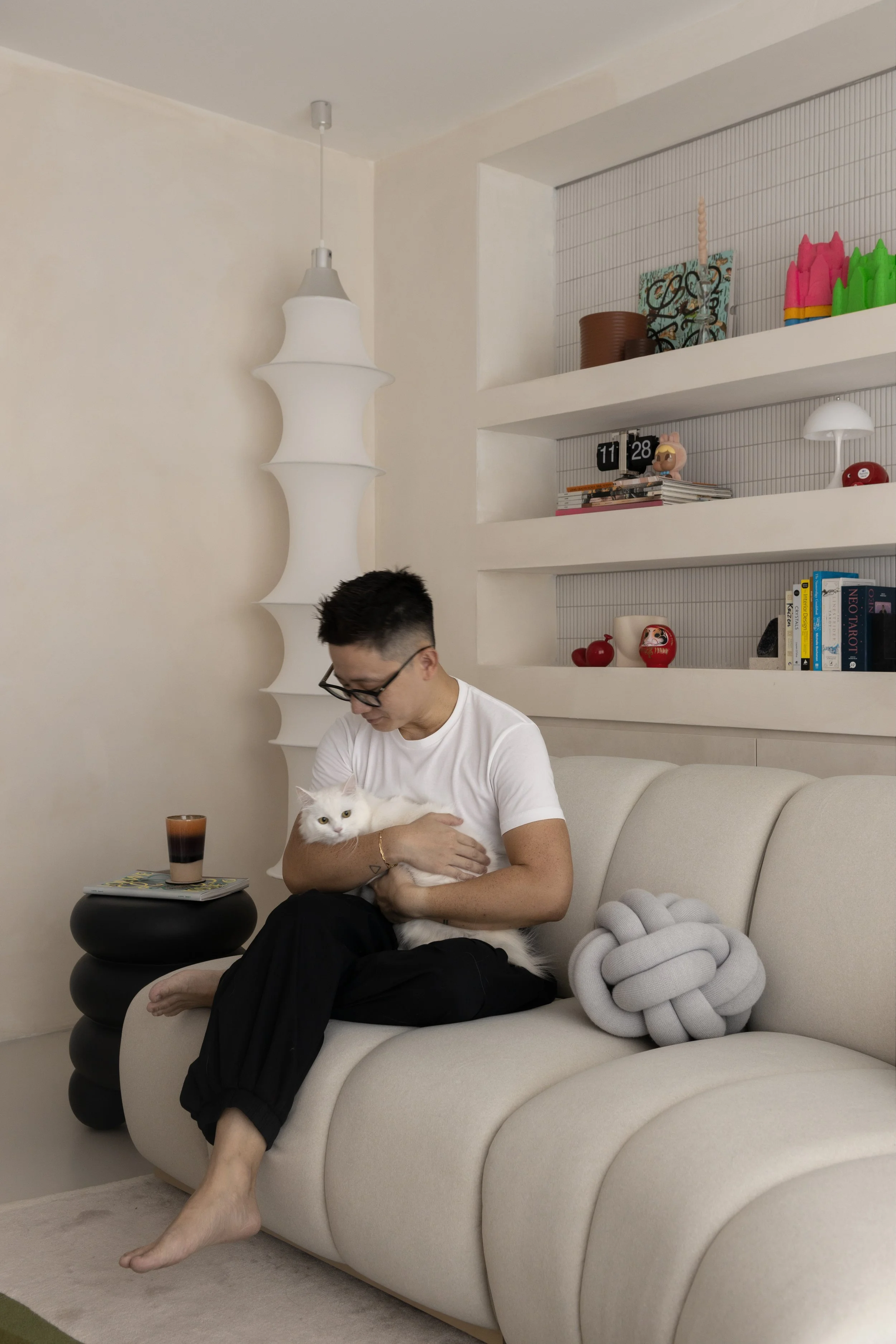 A man sitting on a beige sofa holding a white cat, with a side table with a glass of drink, and shelves with decorative items in the background.