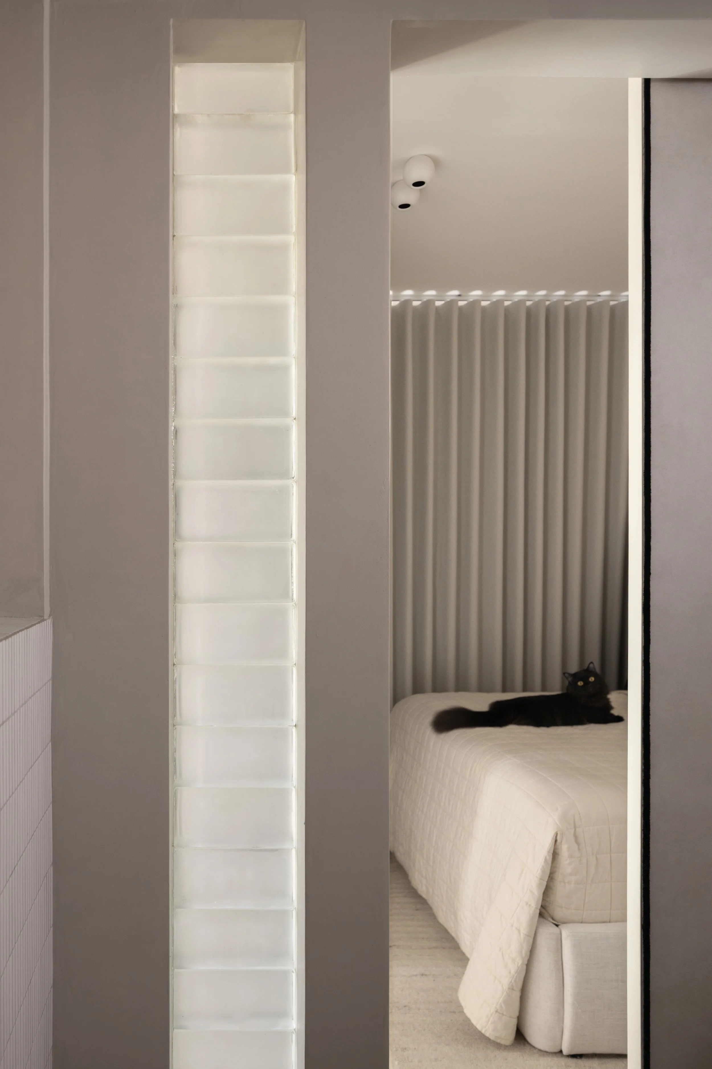 A bedroom with a black cat lying on a white bed near the headboard, partially visible through an open sliding door, with vertical striped curtains in the background and a ceiling light.