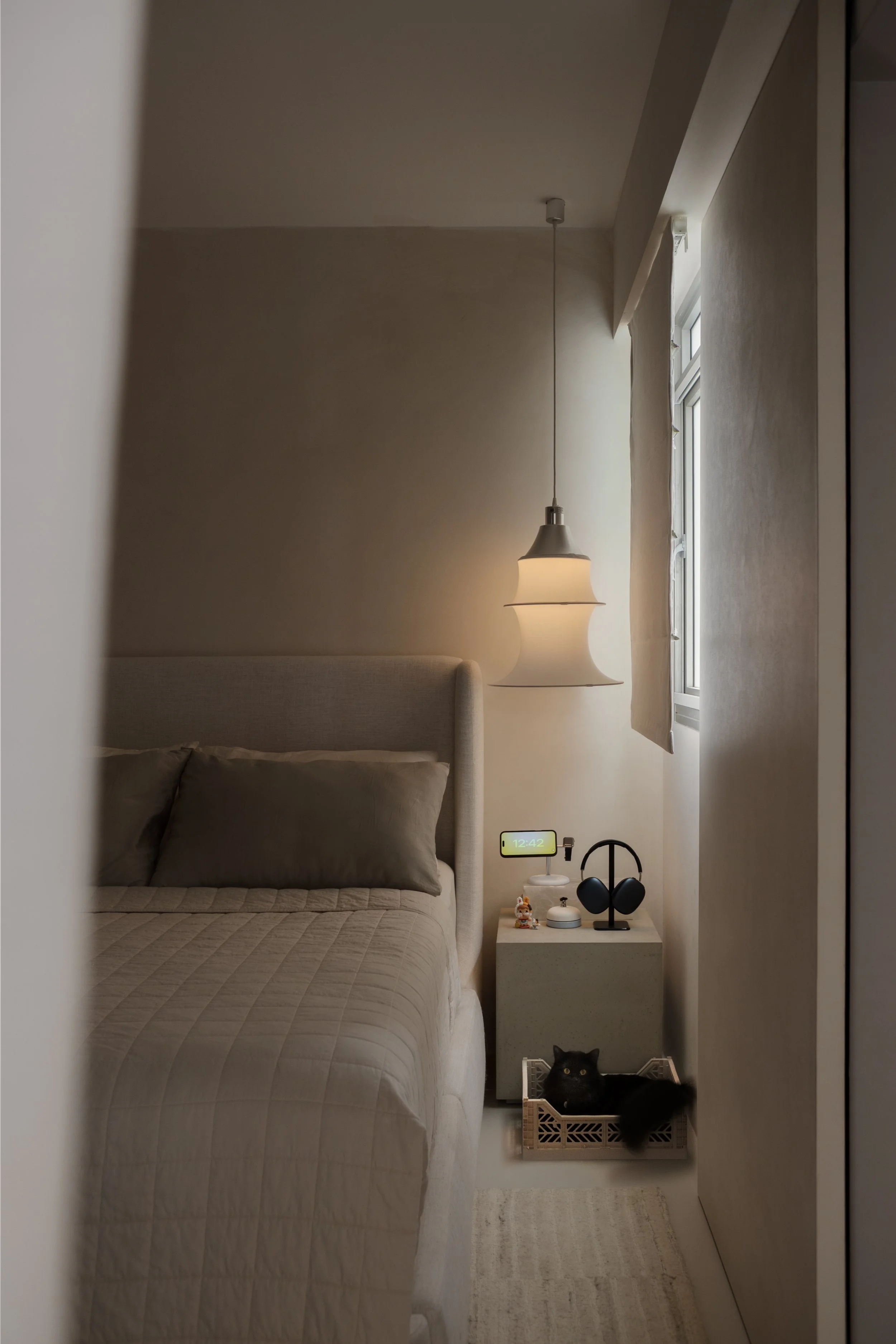 A cozy bedroom corner with a bed and a nightstand. The nightstand has a digital alarm clock, a white lamp, a small figurine, black headphones, and some other small objects. There is a window with beige curtains and a black cat sitting in a basket on 