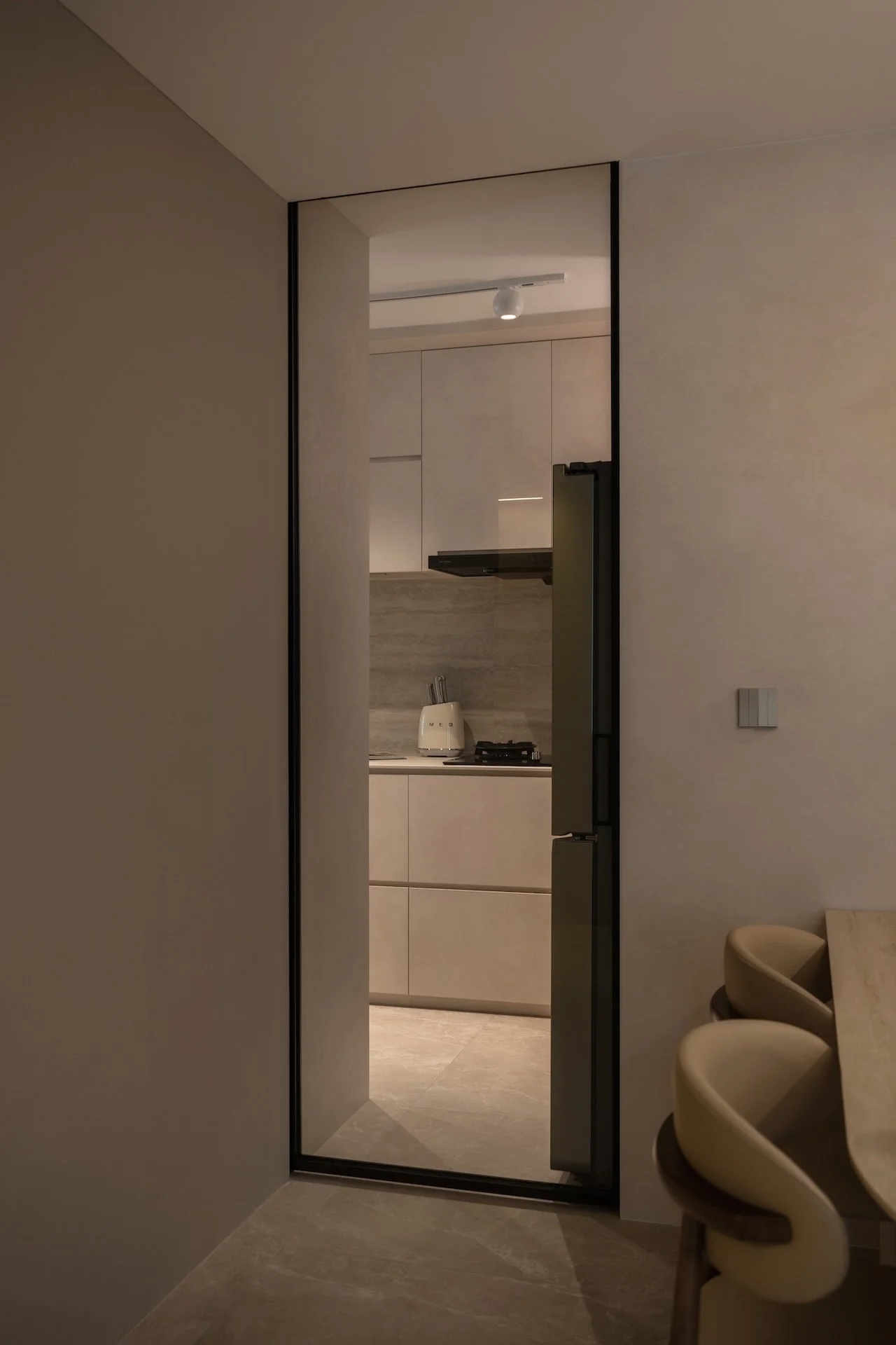 A view into a modern, minimalist kitchen through an open door with a glass panel, featuring beige cabinets, a white kettle, a stovetop, and sleek design elements.