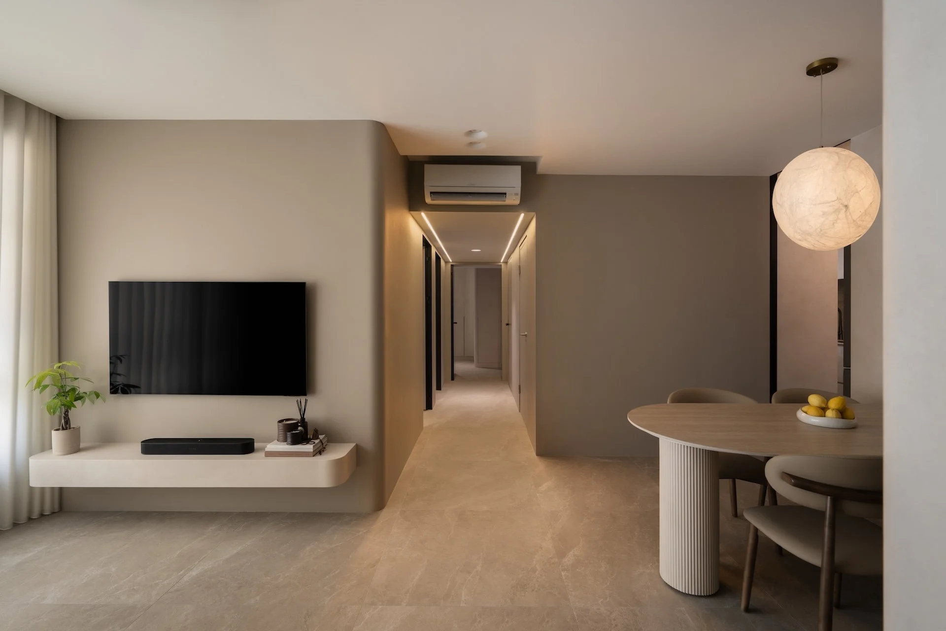 Modern living room with a wall-mounted TV, beige walls, a hallway leading to other rooms, a dining table with chairs, and a round hanging light fixture.