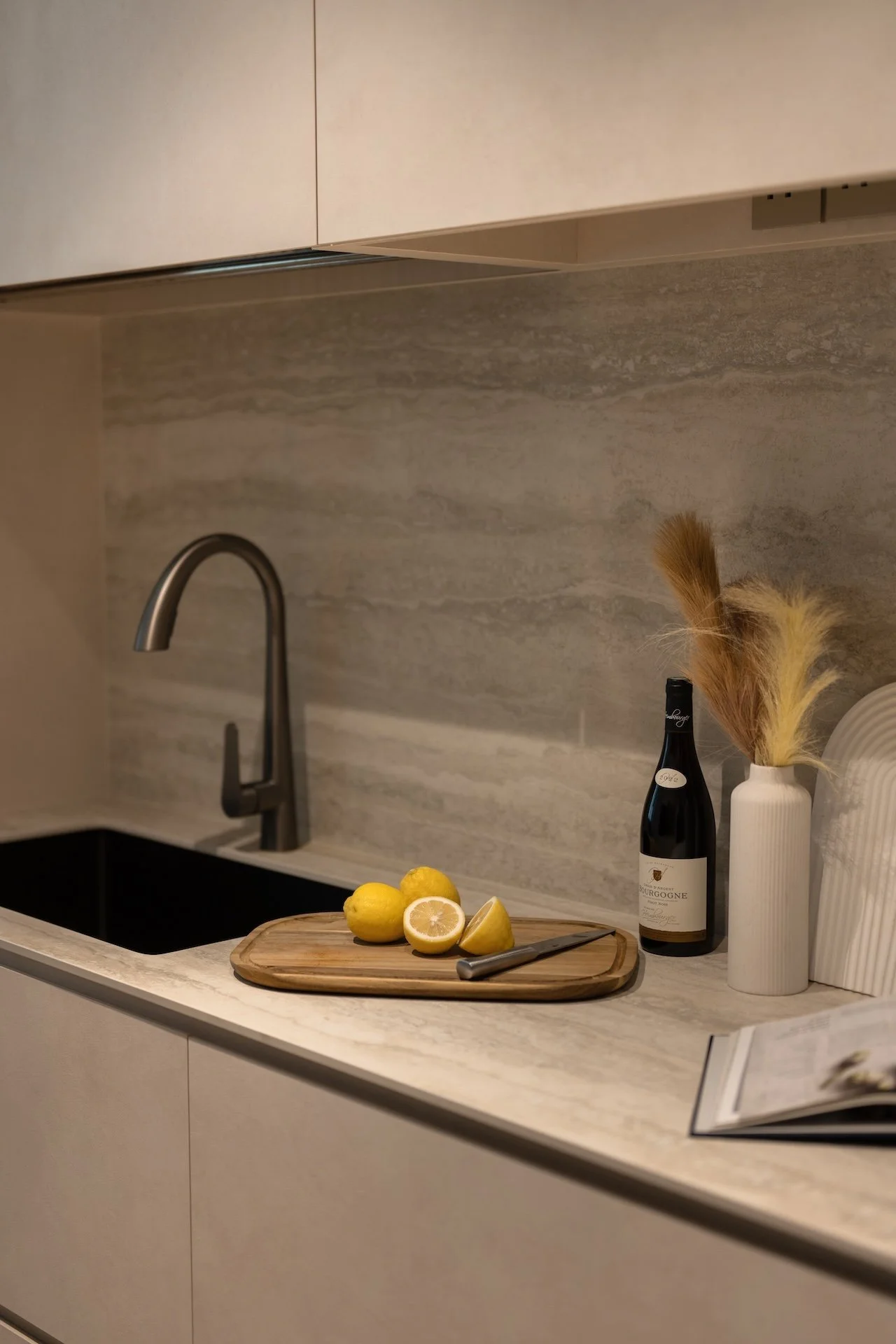 A kitchen countertop with a lemon, a lemon squeezer, a bottle of wine, a toilet paper roll, and a white vase with dried grasses.